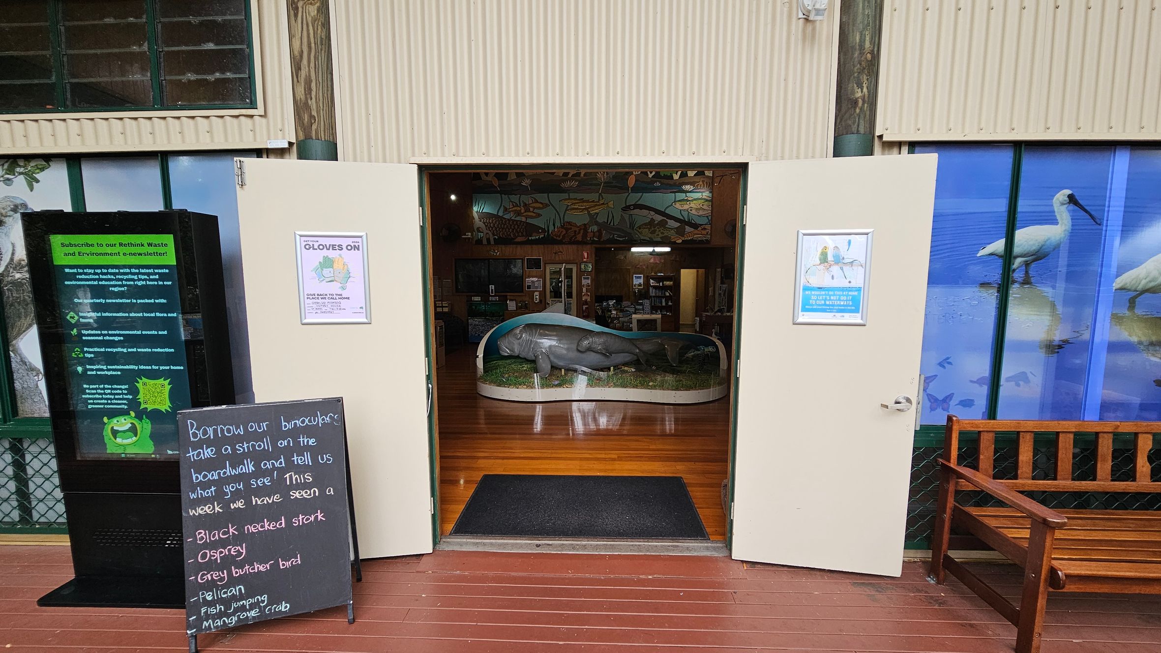 Open double doors lead into a room with a large manatee model displayed on a platform in the centre. The floor is wooden, and the walls feature colourful aquatic-themed murals. To the left of the entrance, a blackboard lists wildlife sightings, and a digital sign promotes an environmental newsletter. On the right, a wooden bench sits in front of a window with images of birds. The ceiling is not visible.