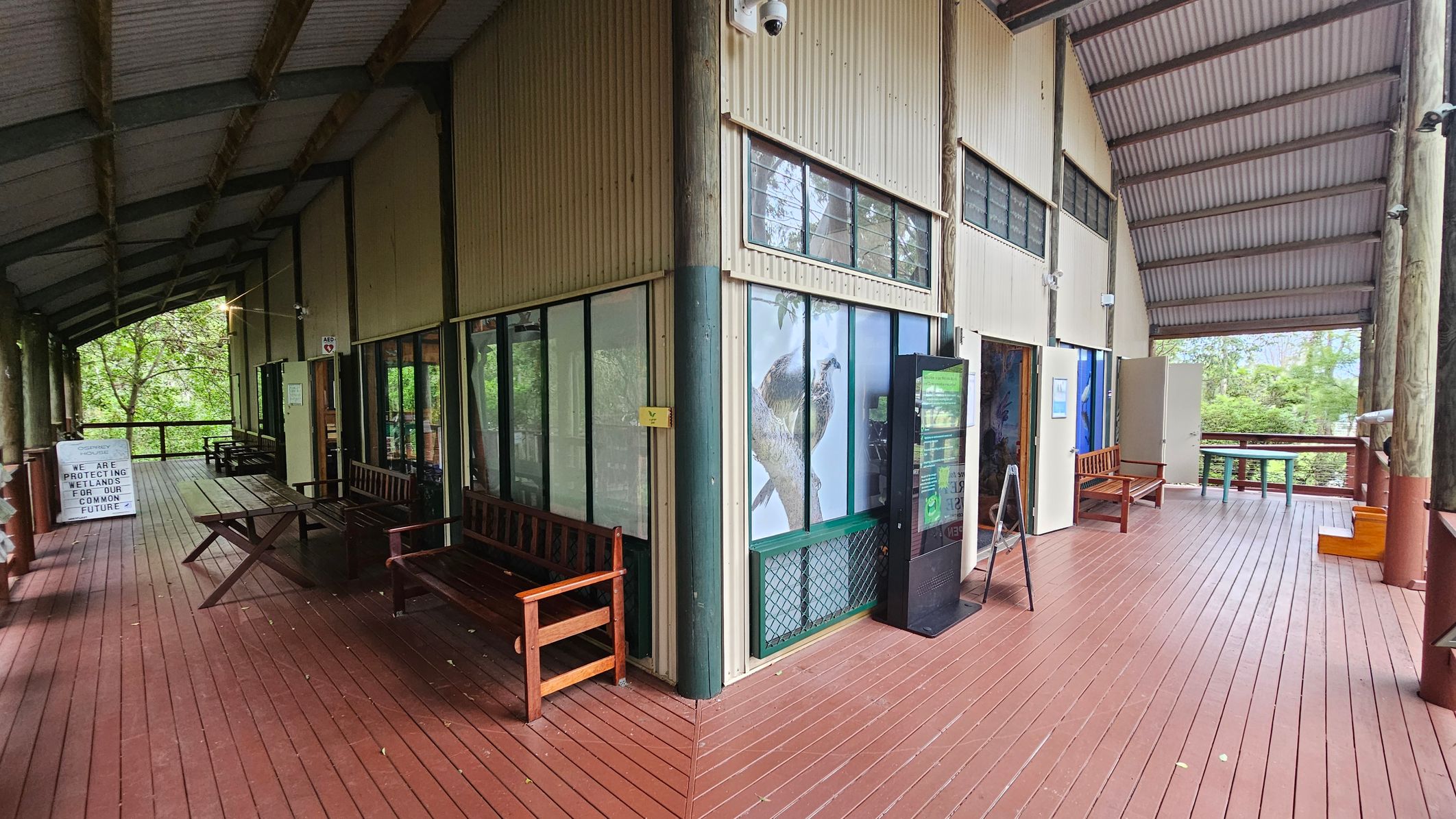 A covered wooden deck with a high, corrugated metal ceiling. On the left, there are wooden benches and a sign about wetland protection. The middle section features large windows with a bird image and a digital display. On the right, more benches and a railing overlook greenery. The floor is made of reddish-brown wooden planks.