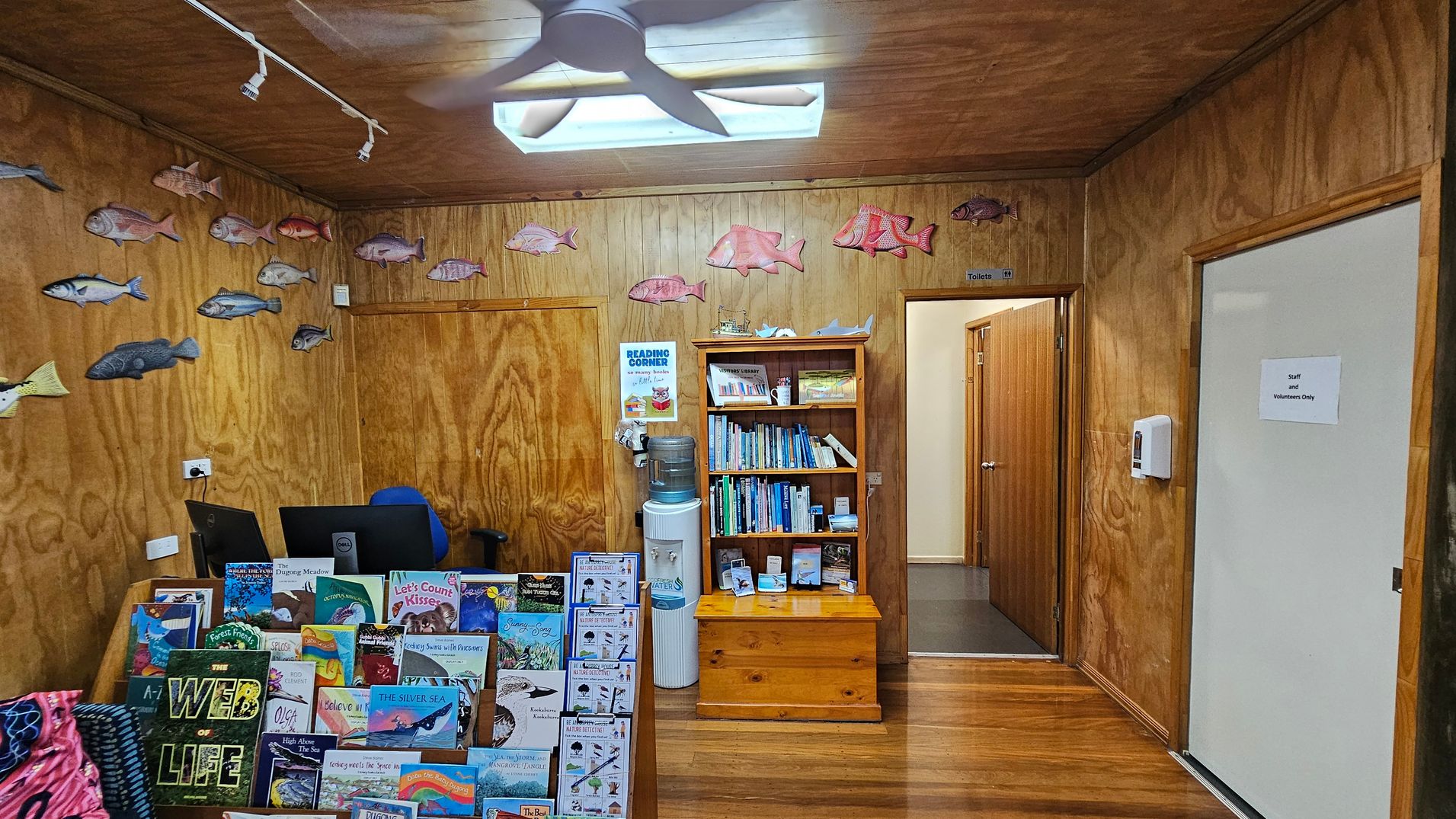 A room with wooden walls and floor. On the left, there are two computer monitors on a desk, with a display of children's books in front. Fish decorations are mounted on the walls. In the centre, a wooden bookshelf filled with books stands next to a water cooler. A sign above the bookshelf reads "Reading Corner." To the right, a door marked "Staff and Volunteers Only" is visible. A ceiling fan and light fixture are above.