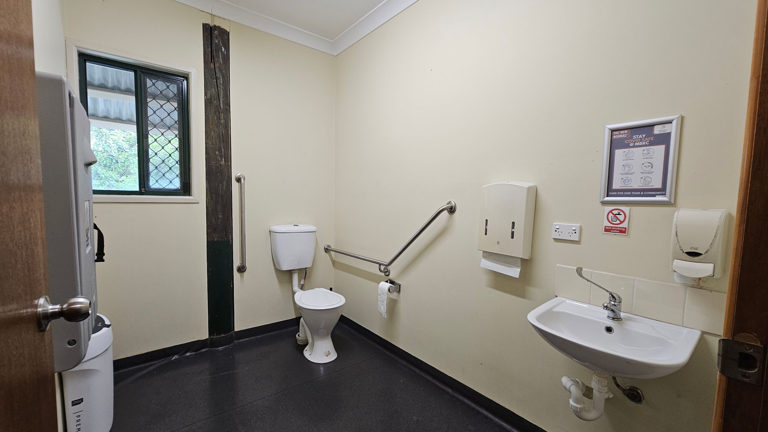 A public restroom with a white toilet and grab bars on the left. A small window with a security grille is above the toilet. On the right, a white sink is mounted on the wall with a soap dispenser and paper towel holder nearby. A framed sign and a no-smoking notice are above the sink. The floor is dark and the walls are light-coloured. The ceiling is white with a simple design.