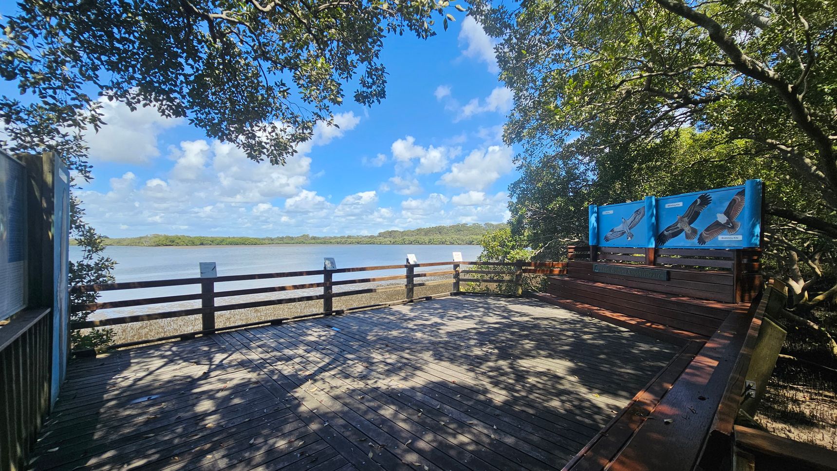 A wooden viewing platform with railings overlooks a body of water surrounded by trees. On the right, there are informational signs featuring images of birds. The platform is partially shaded by overhanging branches. The floor is made of wooden planks, and the background shows a clear view of the water and distant tree line.