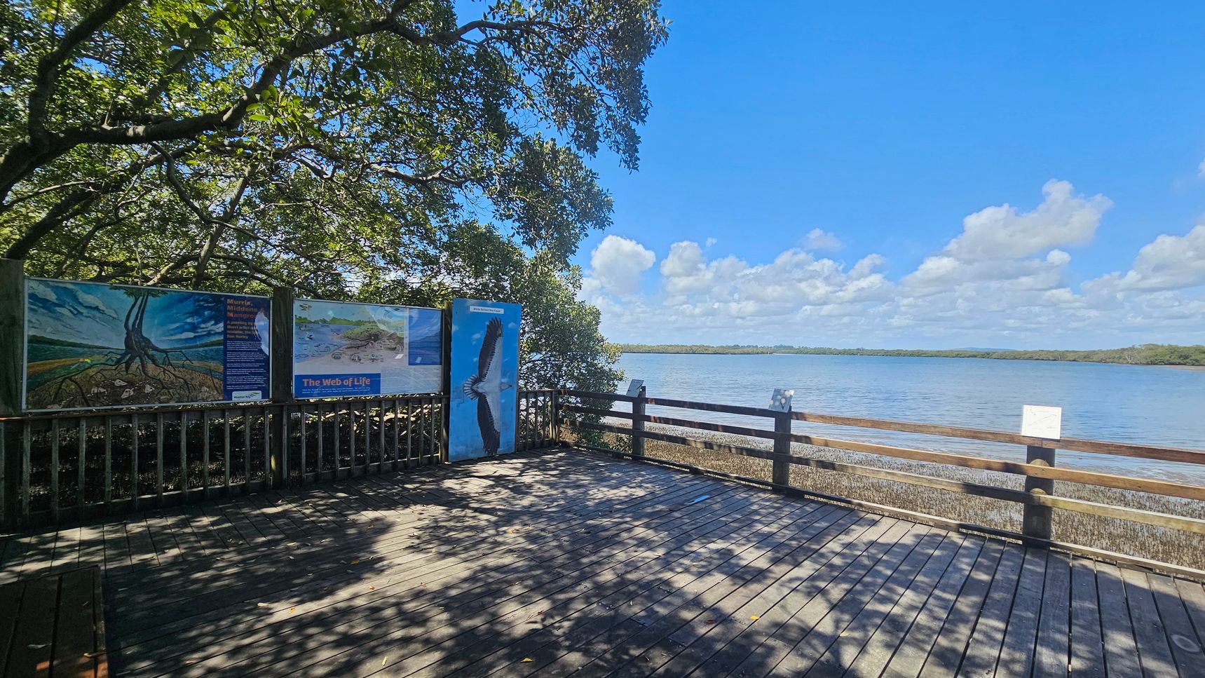 A wooden deck overlooks a body of water with a railing on the right. On the left, there are informational signs with images, including one of a bird. Trees with dense foliage provide shade, casting shadows on the deck. The background features a calm water surface extending to a distant tree-lined shore under a blue sky with scattered clouds.