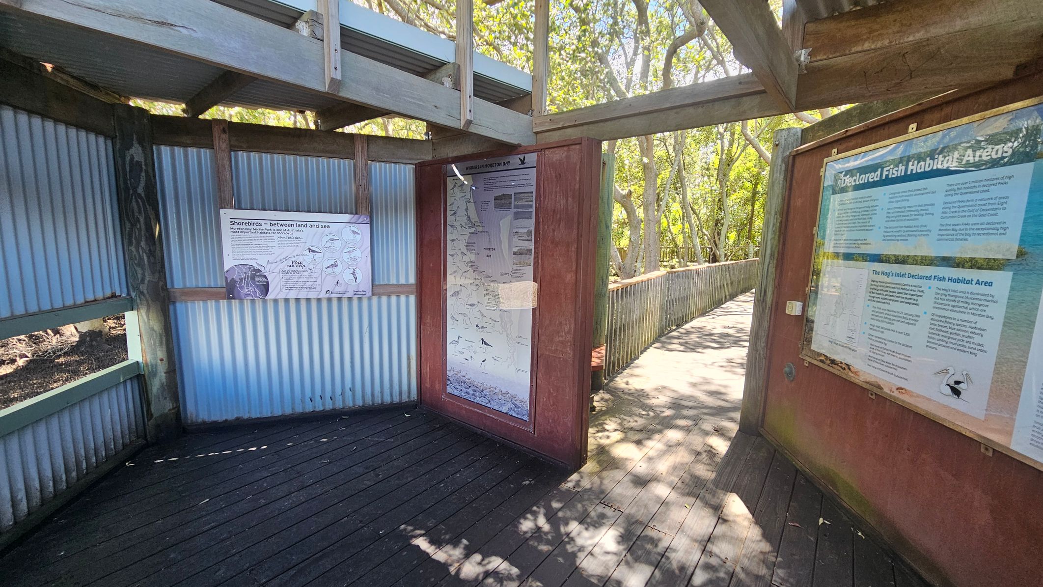 A wooden walkway with corrugated metal walls on the left, featuring informational signs about shorebirds and fish habitats. The floor is made of dark wooden planks. On the right, a large sign provides details about declared fish habitat areas. The walkway extends into a wooded area with trees and dappled sunlight filtering through the leaves. The ceiling is partially open with wooden beams.
