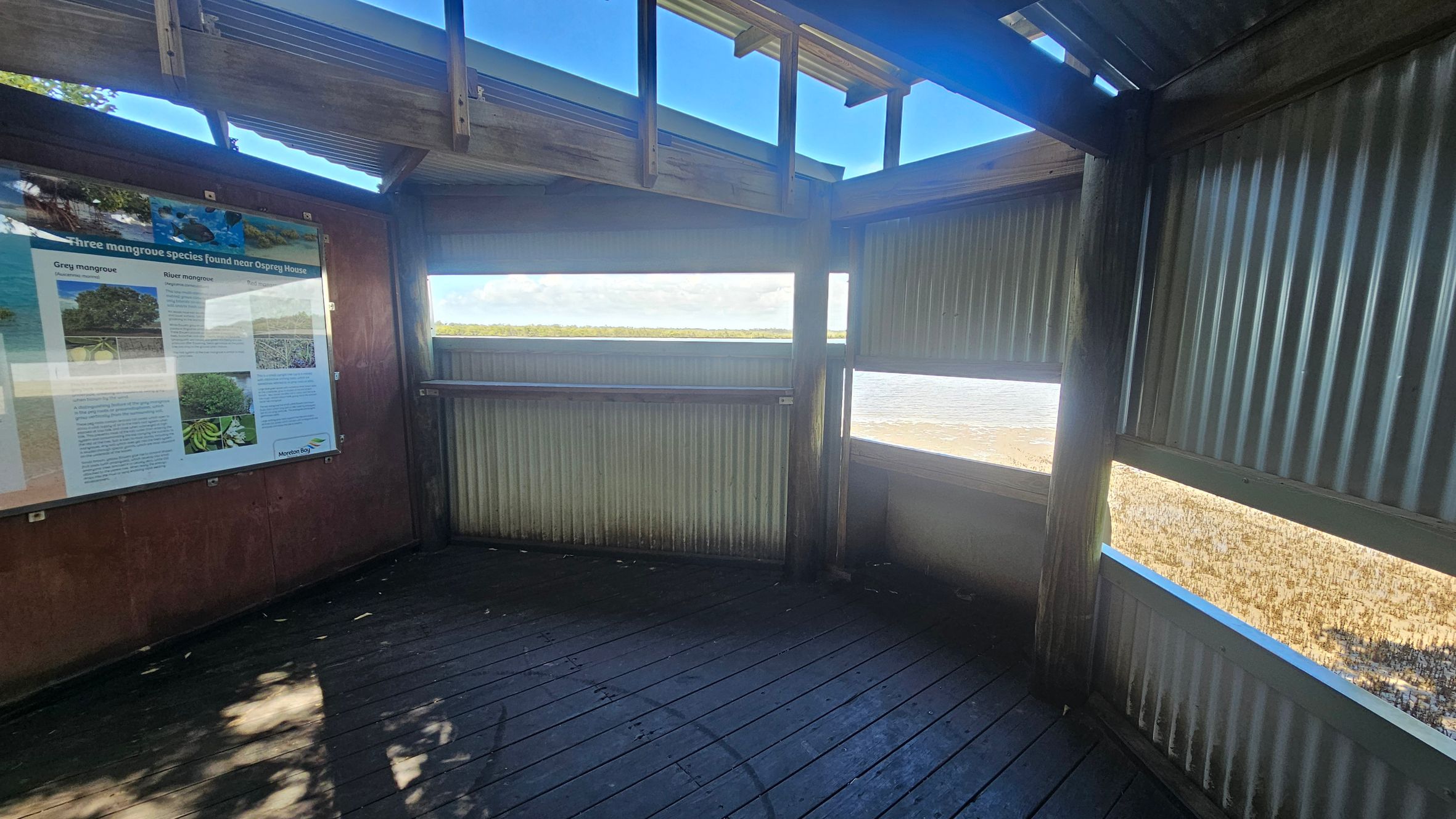 A wooden observation structure with corrugated metal walls and a slatted roof. On the left, a large informational sign about mangrove species is mounted on the wall. The middle and right sections have horizontal openings offering views of a water body and distant greenery. The floor is made of dark wooden planks, and sunlight casts shadows inside.