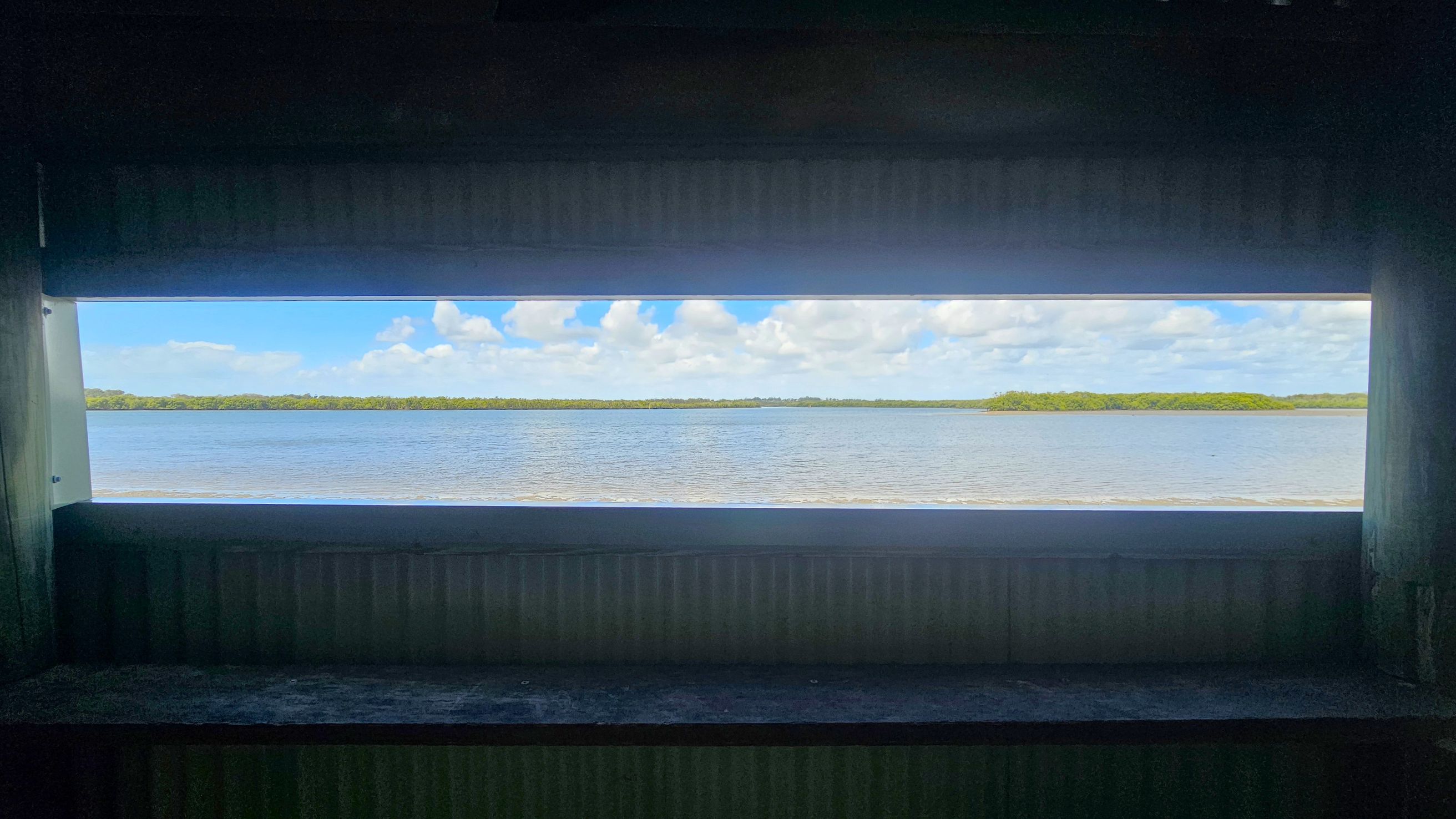 A rectangular window view shows a calm body of water stretching across the middle, with a distant tree-lined shore on the horizon. The foreground is dark, with a ledge beneath the window. The walls surrounding the window are also dark, creating a frame for the bright outdoor scene.