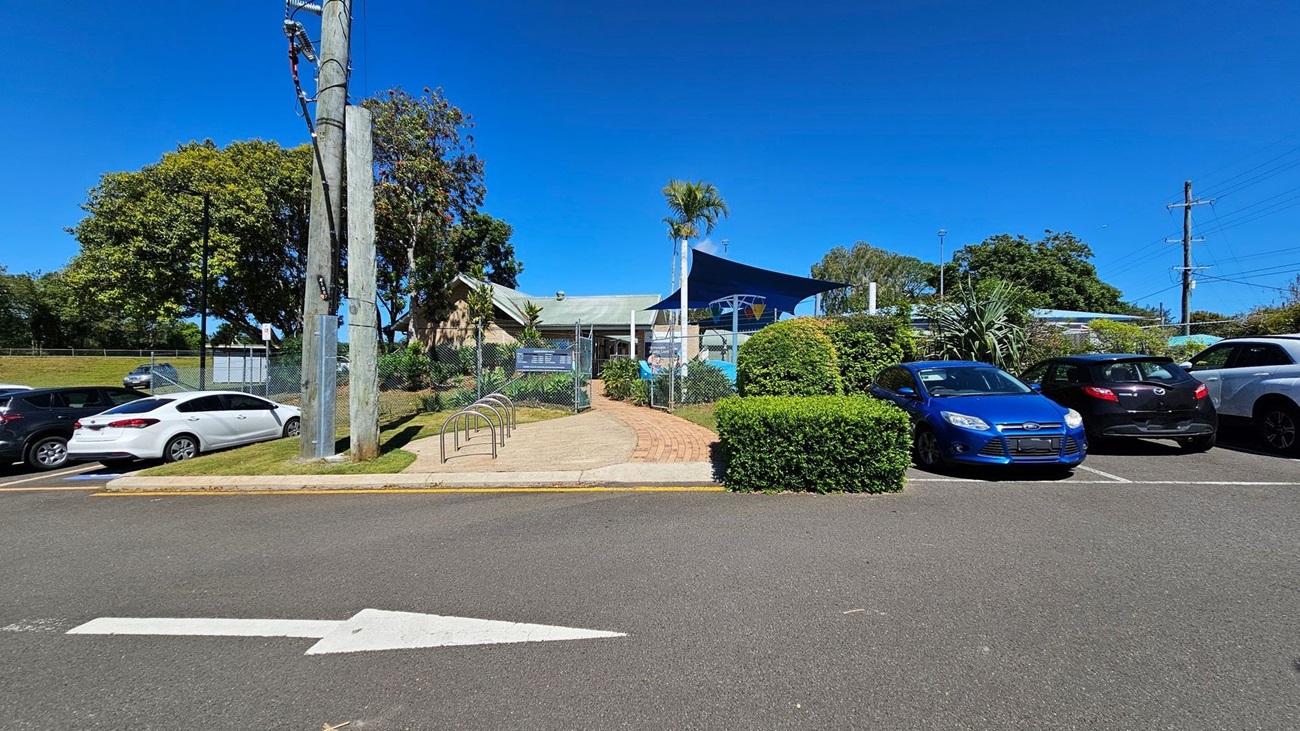 A paved pathway leads to a building with a green roof and a blue shade structure. The entrance is flanked by a chain-link fence and greenery. On the left, there is a row of parked cars, including a white car. On the right, there are more parked cars, including a blue car and a black car. A large utility pole stands near the centre. The background features trees and additional greenery. The foreground shows an asphalt road with a white arrow pointing forward.