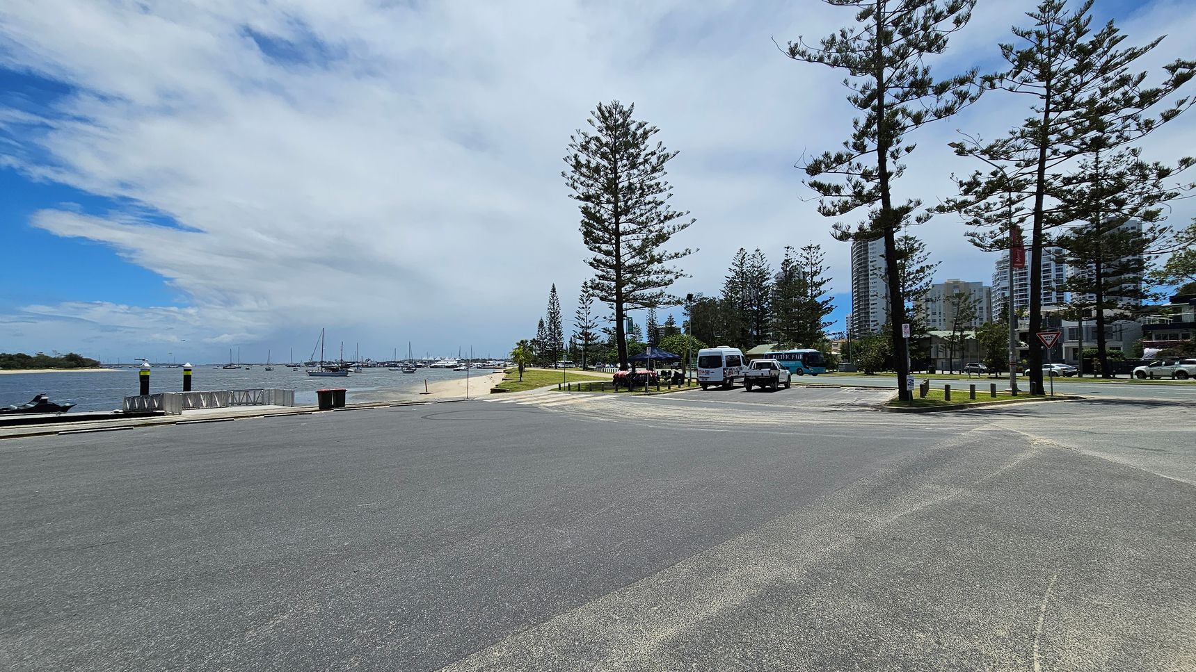 A coastal scene with a wide road in the foreground. On the left, a body of water with several moored sailboats and a small sandy beach. In the middle, tall pine trees line the grassy area, with a few parked vehicles, including a van and a bus. On the right, modern high-rise buildings are visible behind more trees. The sky is partly cloudy, with patches of blue visible. A coastal scene with a wide road in the foreground. On the left, a body of water with several moored sailboats and a small sandy beach. In the middle, tall pine trees line the grassy area, with a few parked vehicles, including a van and a bus. On the right, modern high-rise buildings are visible behind more trees. The sky is partly cloudy, with patches of blue visible.