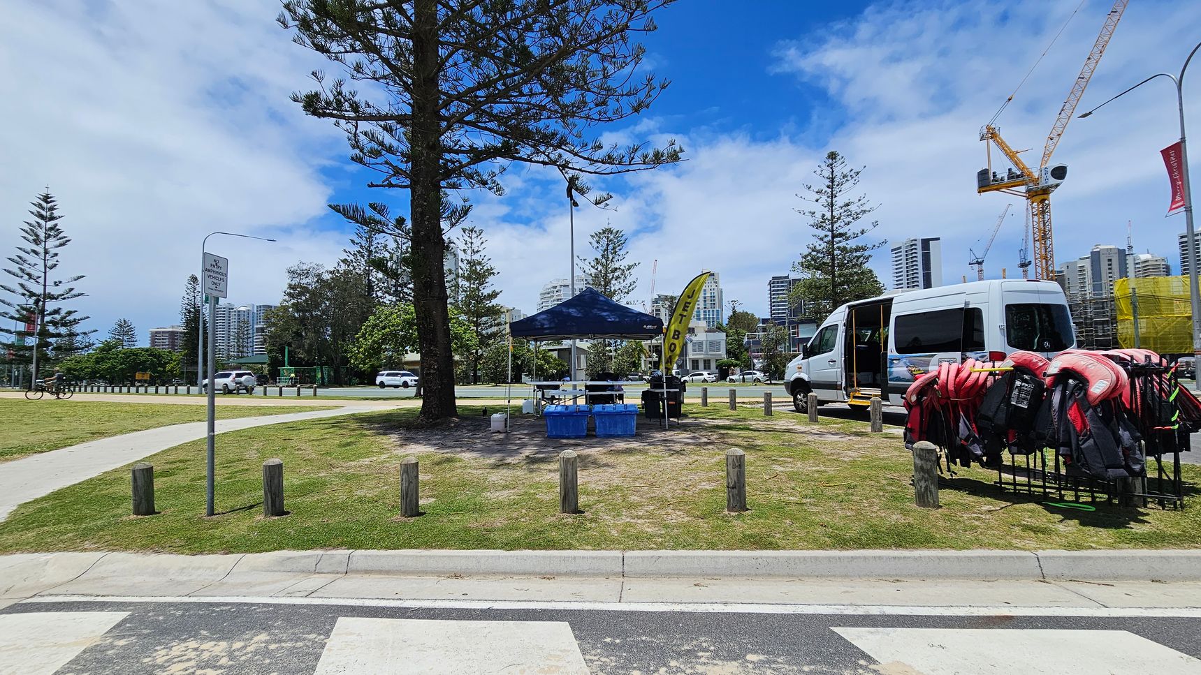 A park area with a small blue tent in the centre, surrounded by trees. To the right, a white van is parked with its door open, and a rack of life jackets is visible. A yellow flag with text is near the tent. In the background, there are tall buildings and a construction crane. A cyclist is on the left, and a signpost is visible. The ground is grassy with a paved path, and wooden bollards line the area. A park area with a small blue tent in the centre, surrounded by trees. To the right, a white van is parked with its door open, and a rack of life jackets is visible. A yellow flag with text is near the tent. In the background, there are tall buildings and a construction crane. A cyclist is on the left, and a signpost is visible. The ground is grassy with a paved path, and wooden bollards line the area.