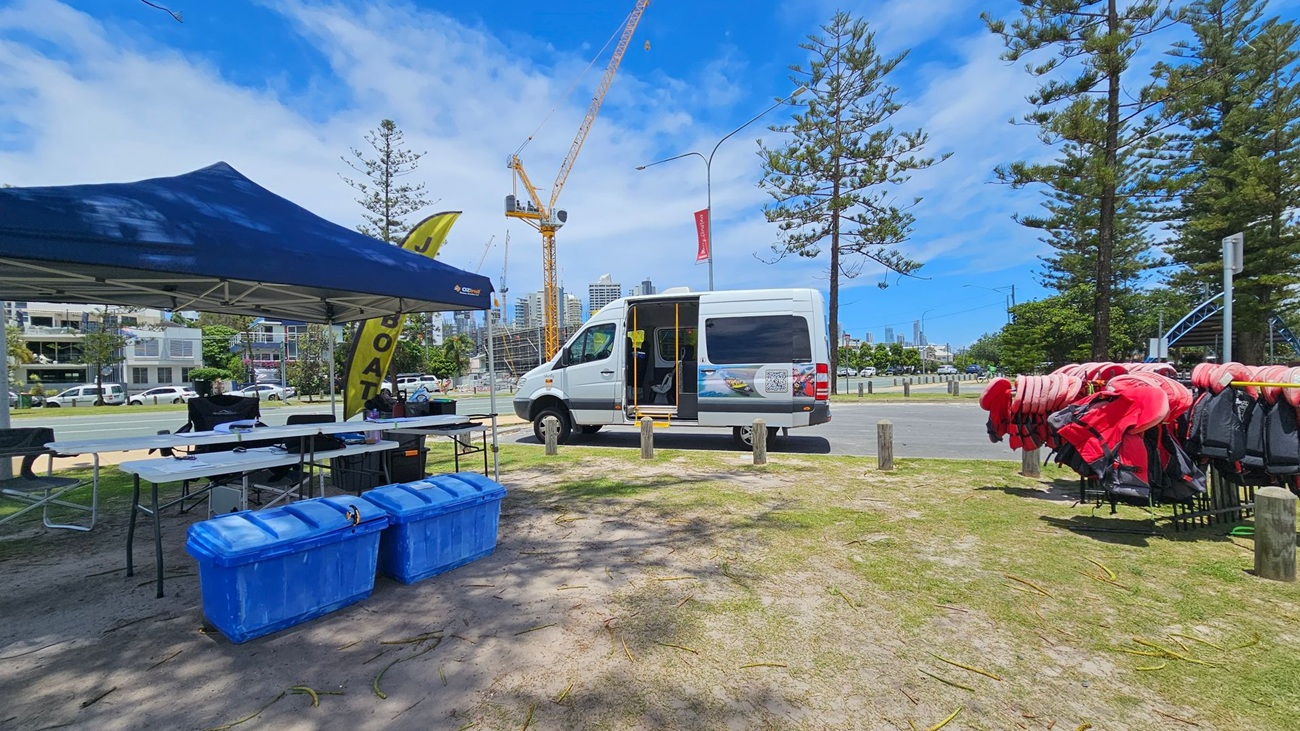 On the left, a blue canopy tent covers tables with various items and two large blue storage bins. In the centre, a white van with its side door open is parked on a grassy area. To the right, a rack holds numerous red life jackets. In the background, there are tall trees, a construction crane, and buildings. The ground is a mix of grass and sand, and a road with parked cars runs behind the setup. On the left, a blue canopy tent covers tables with various items and two large blue storage bins. In the centre, a white van with its side door open is parked on a grassy area. To the right, a rack holds numerous red life jackets. In the background, there are tall trees, a construction crane, and buildings. The ground is a mix of grass and sand, and a road with parked cars runs behind the setup.