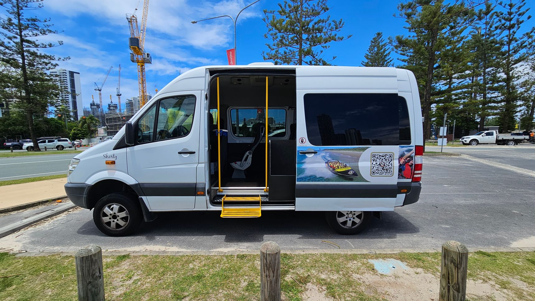A white van with an open sliding door and yellow steps is parked on a paved area. The van has a graphic of a speedboat and a QR code on its side. In the background, there are tall pine trees and several construction cranes near high-rise buildings. A few vehicles are visible on the road, and the ground is a mix of grass and sand. A white van with an open sliding door and yellow steps is parked on a paved area. The van has a graphic of a speedboat and a QR code on its side. In the background, there are tall pine trees and several construction cranes near high-rise buildings. A few vehicles are visible on the road, and the ground is a mix of grass and sand.