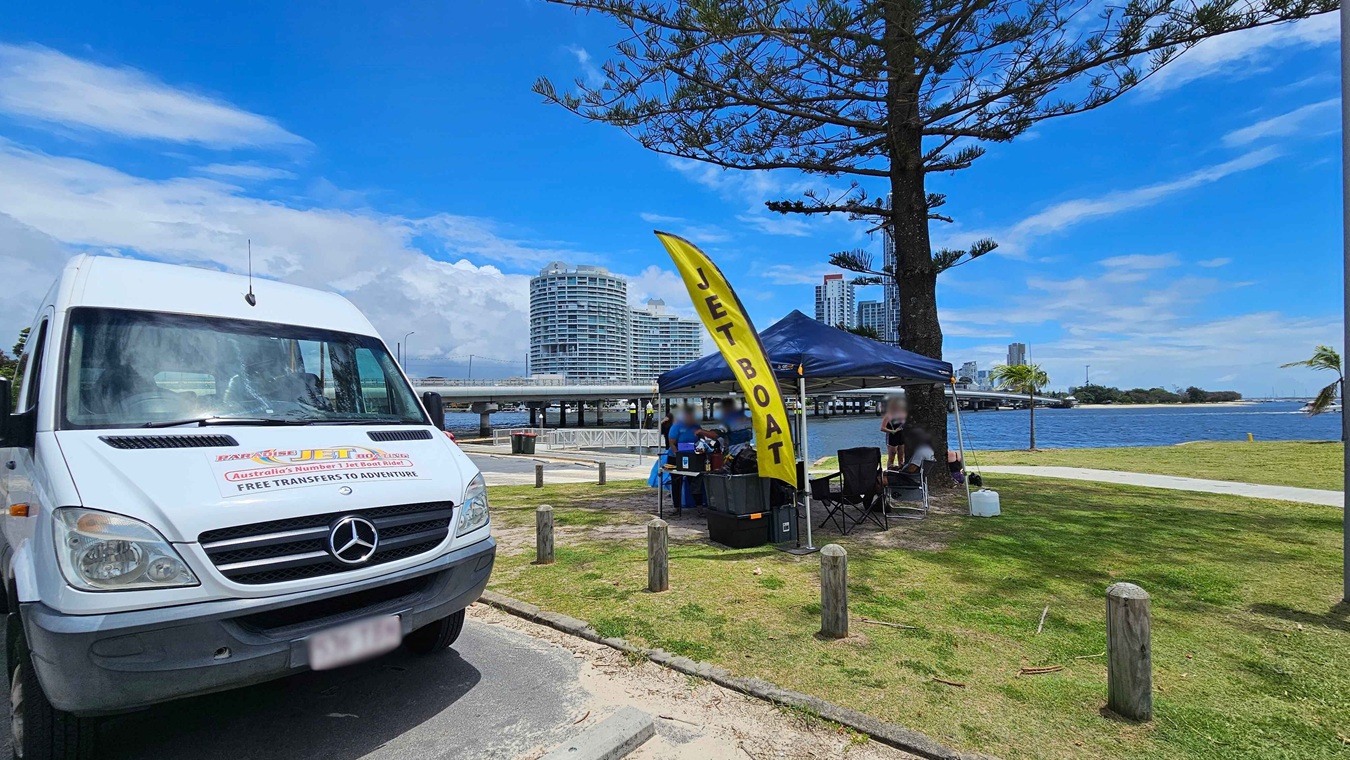 A white van with a Mercedes logo is parked on the left, featuring signage for jet boat rides. On the right, a yellow flag with "JET BOAT" written on it stands next to a blue canopy tent. Under the tent, people are gathered around a table. In the background, a river is visible with a bridge and modern buildings. The area is grassy with a few trees, and wooden posts line the path. A white van with a Mercedes logo is parked on the left, featuring signage for jet boat rides. On the right, a yellow flag with "JET BOAT" written on it stands next to a blue canopy tent. Under the tent, people are gathered around a table. In the background, a river is visible with a bridge and modern buildings. The area is grassy with a few trees, and wooden posts line the path.