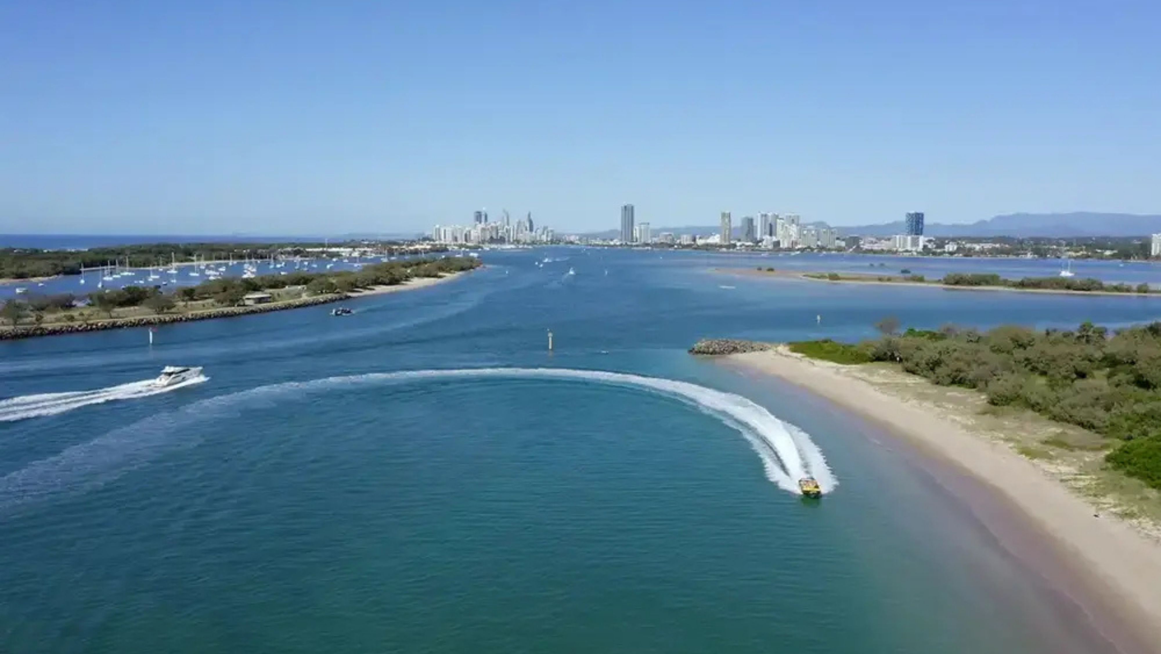 A yellow speedboat with passengers is moving swiftly along the water near a sandy beach. The boat creates a large spray of water as it speeds by. On the left, the water is a deep blue, while on the right, the sandy beach stretches out with some greenery and trees in the background. A rocky breakwater is visible in the distance on the left side. A yellow speedboat with passengers is moving swiftly along the water near a sandy beach. The boat creates a large spray of water as it speeds by. On the left, the water is a deep blue, while on the right, the sandy beach stretches out with some greenery and trees in the background. A rocky breakwater is visible in the distance on the left side.