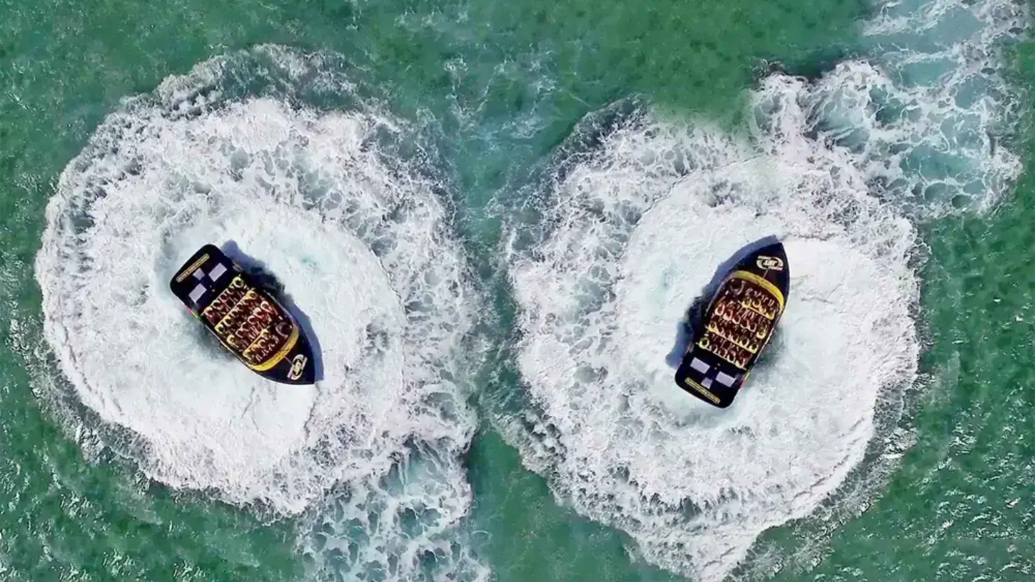Two speedboats are captured from above, each creating circular patterns in the water as they spin. The boats are black with yellow accents and are filled with passengers wearing life jackets. The water is a clear turquoise, and the wake from the boats forms distinct white swirls around them. The scene is dynamic, with the boats positioned side by side, each in the centre of its own circular wake. Two speedboats are captured from above, each creating circular patterns in the water as they spin. The boats are black with yellow accents and are filled with passengers wearing life jackets. The water is a clear turquoise, and the wake from the boats forms distinct white swirls around them. The scene is dynamic, with the boats positioned side by side, each in the centre of its own circular wake.