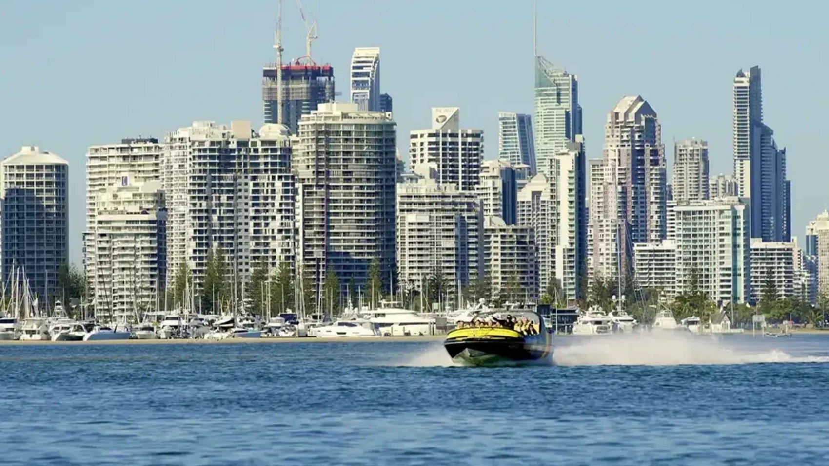 A speedboat moves swiftly across the water in the foreground, creating a trail of spray behind it. In the background, a dense cluster of modern skyscrapers and high-rise buildings form a city skyline. Numerous yachts and boats are docked along the waterfront, with masts visible. The water is a deep blue, contrasting with the light-coloured buildings. The sky is clear, providing a bright backdrop to the scene. A speedboat moves swiftly across the water in the foreground, creating a trail of spray behind it. In the background, a dense cluster of modern skyscrapers and high-rise buildings form a city skyline. Numerous yachts and boats are docked along the waterfront, with masts visible. The water is a deep blue, contrasting with the light-coloured buildings. The sky is clear, providing a bright backdrop to the scene.