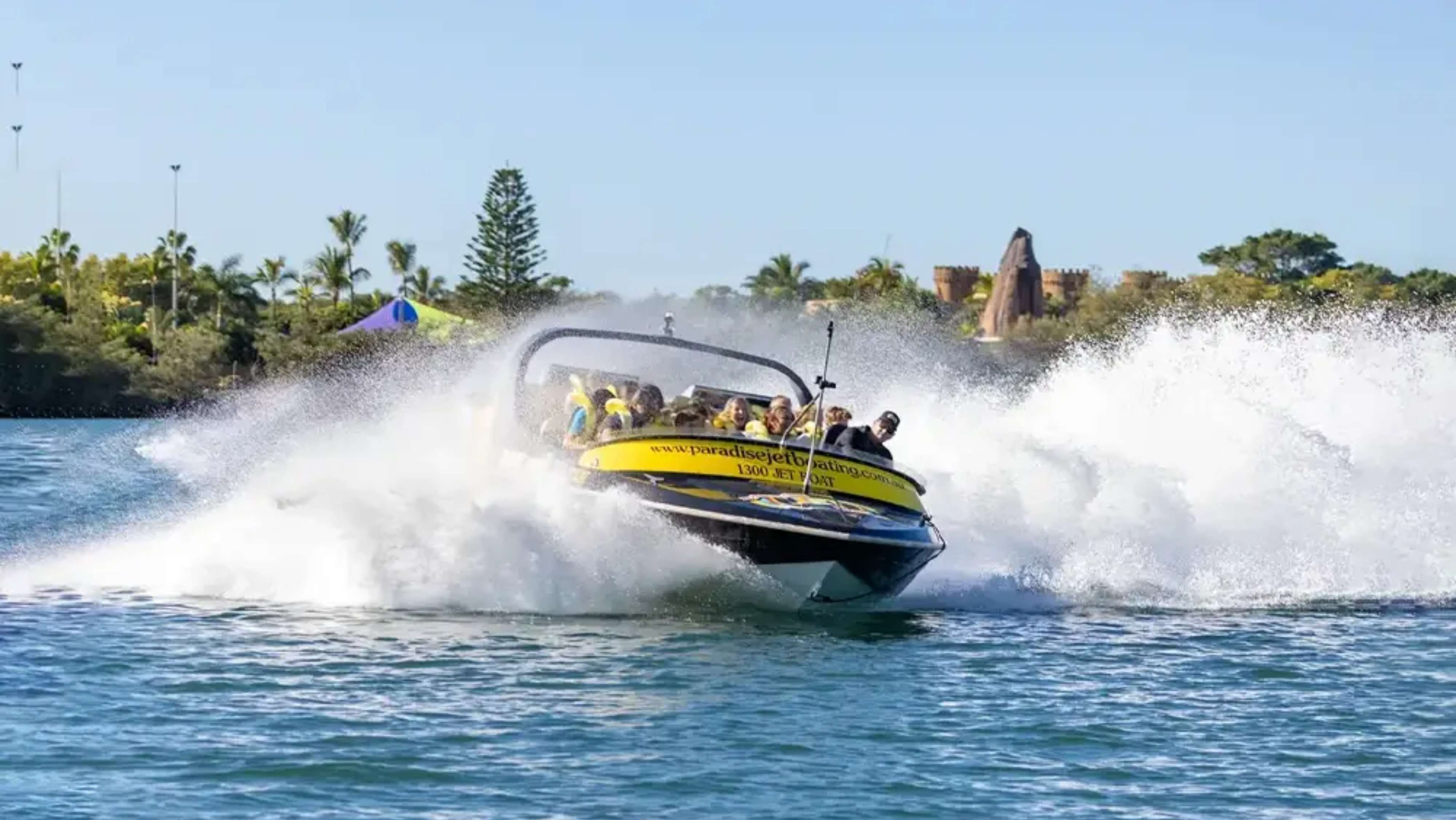 On the left, there are palm trees and a vibrant tent with a blue top, suggesting a recreational area. In the foreground, a jet boat is cutting through the water, creating splashes as it accelerates. The boat has a yellow and black colour scheme, with several passengers visibly enjoying the ride. To the right, there are more trees and what seems to be a decorative stone structure or castle-like feature in the background, possibly part of a theme park. On the left, there are palm trees and a vibrant tent with a blue top, suggesting a recreational area. In the foreground, a jet boat is cutting through the water, creating splashes as it accelerates. The boat has a yellow and black colour scheme, with several passengers visibly enjoying the ride. To the right, there are more trees and what seems to be a decorative stone structure or castle-like feature in the background, possibly part of a theme park.