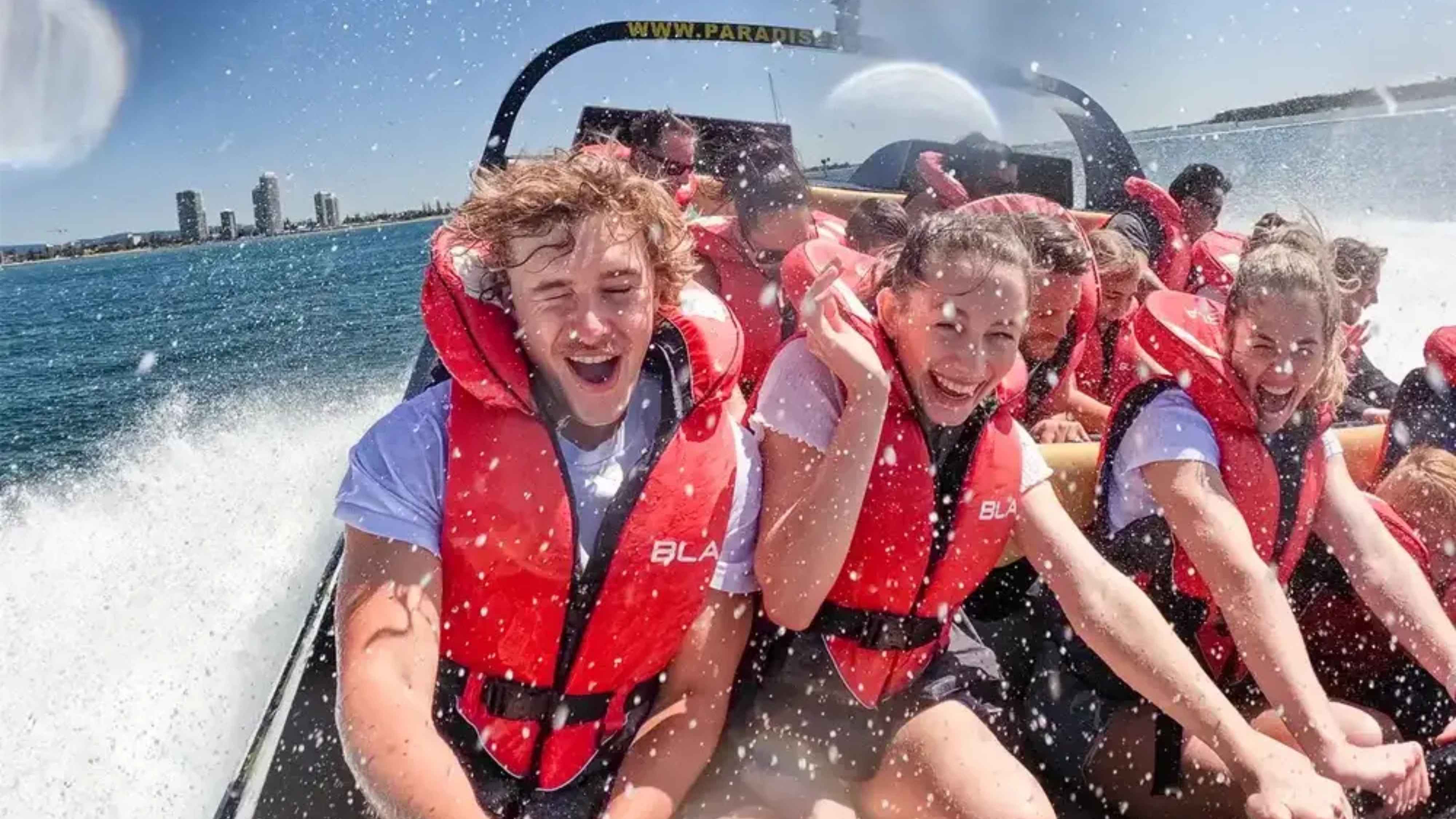 A group of people wearing red life jackets are seated on a speedboat, gripping the safety bars. Water splashes around them, indicating high speed. The background shows a city skyline with several tall buildings against a clear blue sky. The boat's wake is visible on the left, creating a dynamic scene. The people appear to be enjoying the ride, with expressions of excitement. A group of people wearing red life jackets are seated on a speedboat, gripping the safety bars. Water splashes around them, indicating high speed. The background shows a city skyline with several tall buildings against a clear blue sky. The boat's wake is visible on the left, creating a dynamic scene. The people appear to be enjoying the ride, with expressions of excitement.
