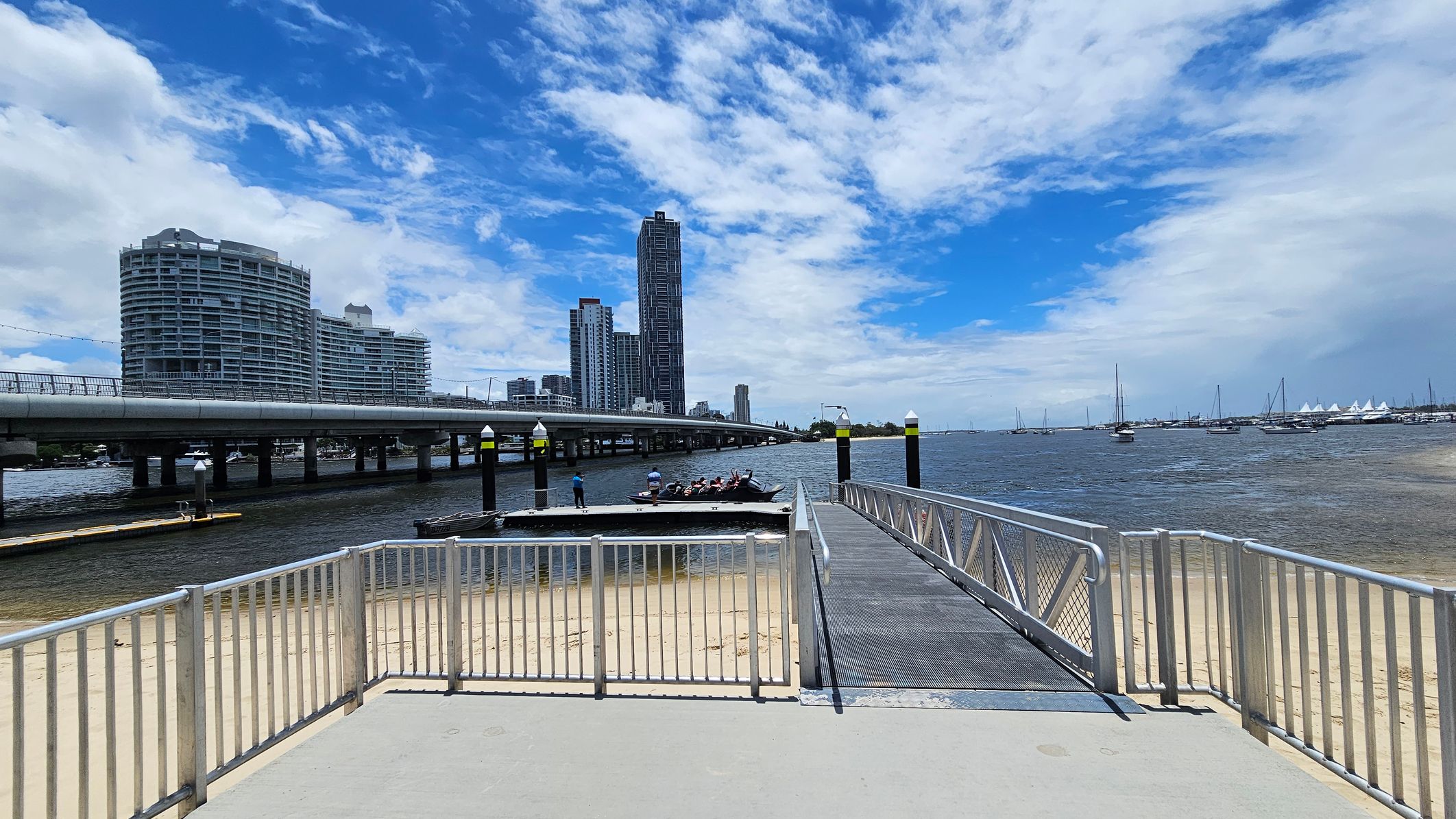 A sandy beach is on the left, with a dock extending into the water. A small boat is moored at the dock. In the background, several sailboats are visible on the water. On the right, there is a paved area with parking spaces and a few tall trees. Beyond the trees, modern high-rise buildings are visible. The scene is calm, with a mix of natural and urban elements. A sandy beach is on the left, with a dock extending into the water. A small boat is moored at the dock. In the background, several sailboats are visible on the water. On the right, there is a paved area with parking spaces and a few tall trees. Beyond the trees, modern high-rise buildings are visible. The scene is calm, with a mix of natural and urban elements.
