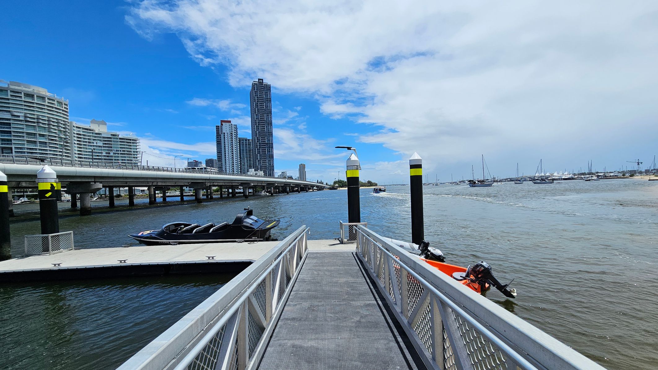 A metal walkway leads to a floating dock on a body of water. On the left, a sleek black boat is moored. In the background, a bridge stretches across the water, with modern high-rise buildings visible beyond it. On the right, several sailboats are anchored in the distance. The water is calm, and the sky is partly cloudy. Two black and yellow posts are positioned near the dock. A metal walkway leads to a floating dock on a body of water. On the left, a sleek black boat is moored. In the background, a bridge stretches across the water, with modern high-rise buildings visible beyond it. On the right, several sailboats are anchored in the distance. The water is calm, and the sky is partly cloudy. Two black and yellow posts are positioned near the dock.