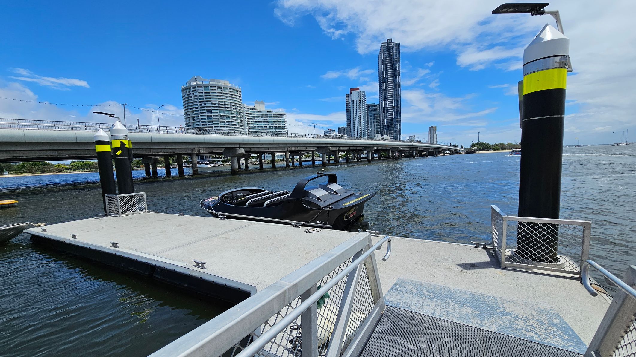 A modern dock with a sleek black boat moored on the right. The dock is bordered by metal railings and has a textured surface. Two tall black and yellow posts with solar panels are positioned on the dock. In the background, a bridge spans across the water, with several high-rise buildings visible, including a cylindrical one on the left and a tall rectangular one on the right. The water is calm, and a few boats are seen in the distance. A modern dock with a sleek black boat moored on the right. The dock is bordered by metal railings and has a textured surface. Two tall black and yellow posts with solar panels are positioned on the dock. In the background, a bridge spans across the water, with several high-rise buildings visible, including a cylindrical one on the left and a tall rectangular one on the right. The water is calm, and a few boats are seen in the distance.