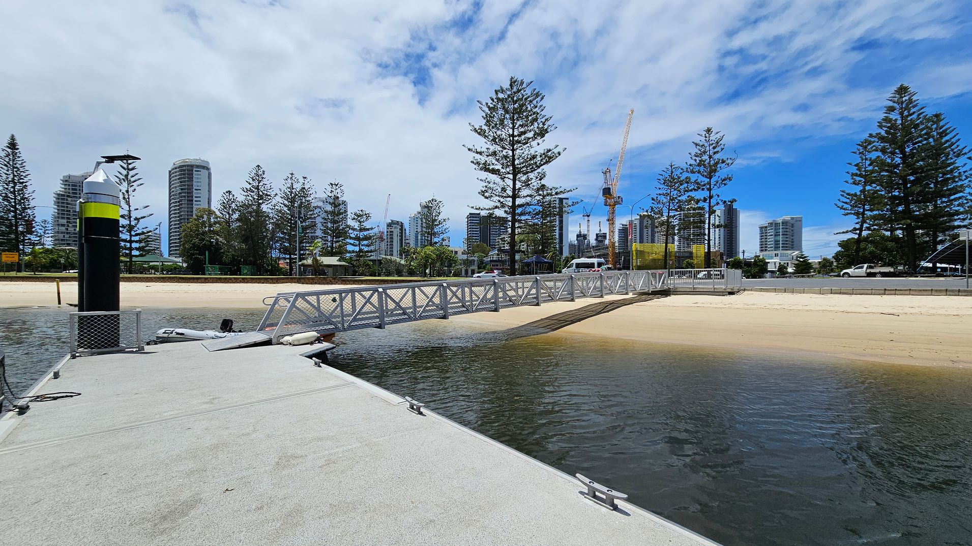 A concrete dock extends into the water on the left, with a black and yellow post at the edge. A metal walkway leads from the dock to a sandy beach on the right. In the background, tall buildings and construction cranes are visible, surrounded by tall pine trees. A road runs parallel to the beach, with a few vehicles on it. The sky is partly cloudy. A concrete dock extends into the water on the left, with a black and yellow post at the edge. A metal walkway leads from the dock to a sandy beach on the right. In the background, tall buildings and construction cranes are visible, surrounded by tall pine trees. A road runs parallel to the beach, with a few vehicles on it. The sky is partly cloudy.