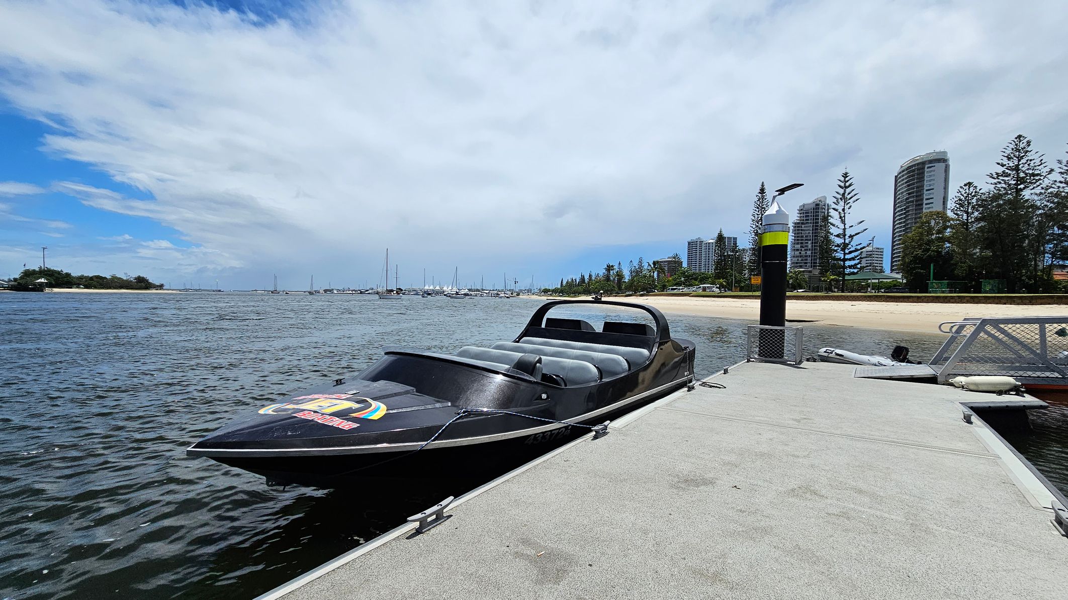 A sleek black speedboat is moored to a concrete dock on the right. The boat has a low profile with grey seating. In the background, a sandy beach is lined with tall pine trees and several modern high-rise buildings. On the left, the water stretches out with numerous sailboats anchored in the distance. A black and yellow striped post is visible on the dock. A sleek black speedboat is moored to a concrete dock on the right. The boat has a low profile with grey seating. In the background, a sandy beach is lined with tall pine trees and several modern high-rise buildings. On the left, the water stretches out with numerous sailboats anchored in the distance. A black and yellow striped post is visible on the dock.