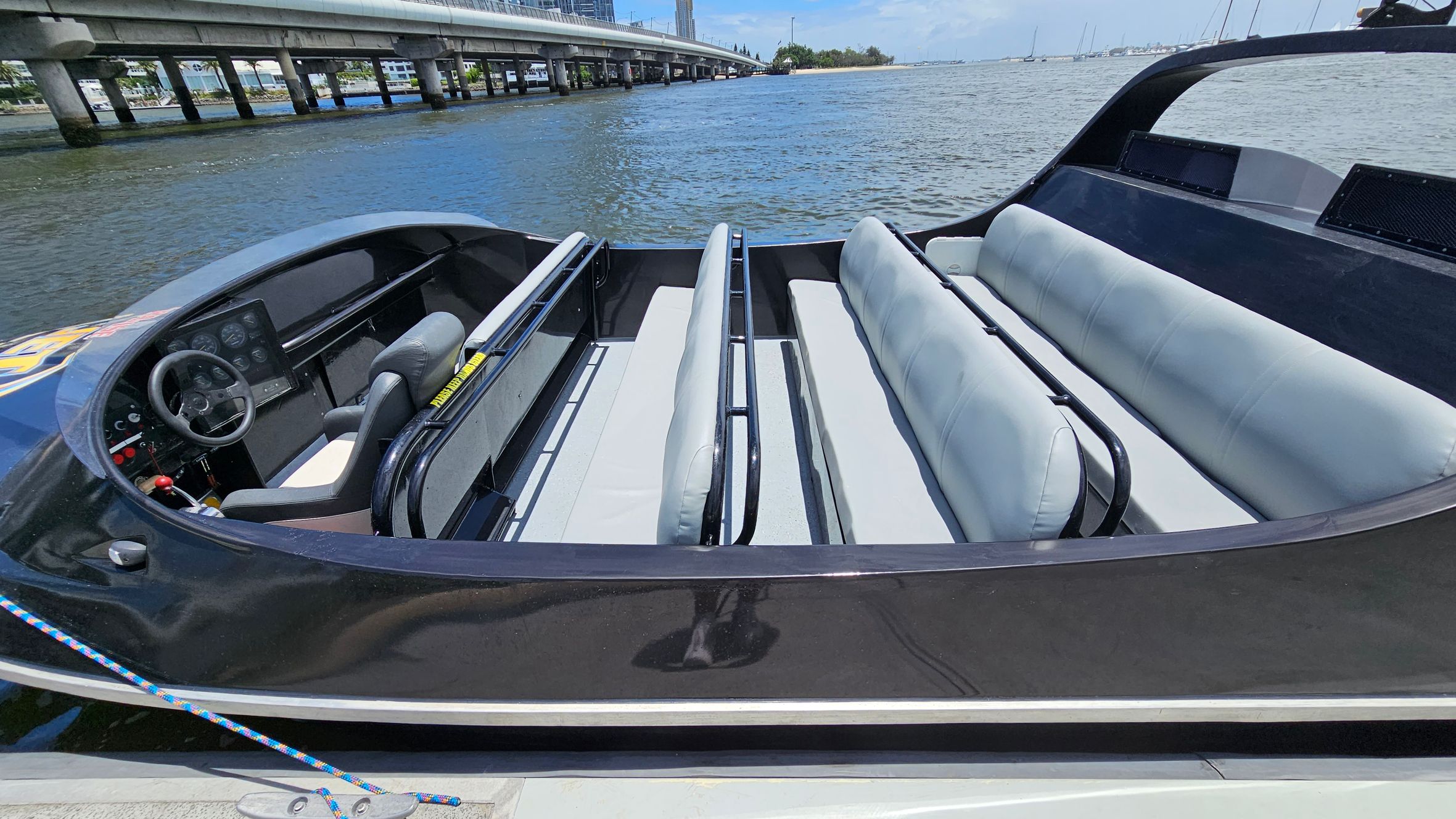A sleek black speedboat with a modern design is docked by the water. The interior features two rows of light grey cushioned seats with black railings. The cockpit area has a steering wheel and a control panel with various gauges and buttons. A colourful rope is tied to the boat's cleat. In the background, a bridge extends over the water, with buildings and trees visible in the distance. The water is calm, and several boats are seen further away. A sleek black speedboat with a modern design is docked by the water. The interior features two rows of light grey cushioned seats with black railings. The cockpit area has a steering wheel and a control panel with various gauges and buttons. A colourful rope is tied to the boat's cleat. In the background, a bridge extends over the water, with buildings and trees visible in the distance. The water is calm, and several boats are seen further away.