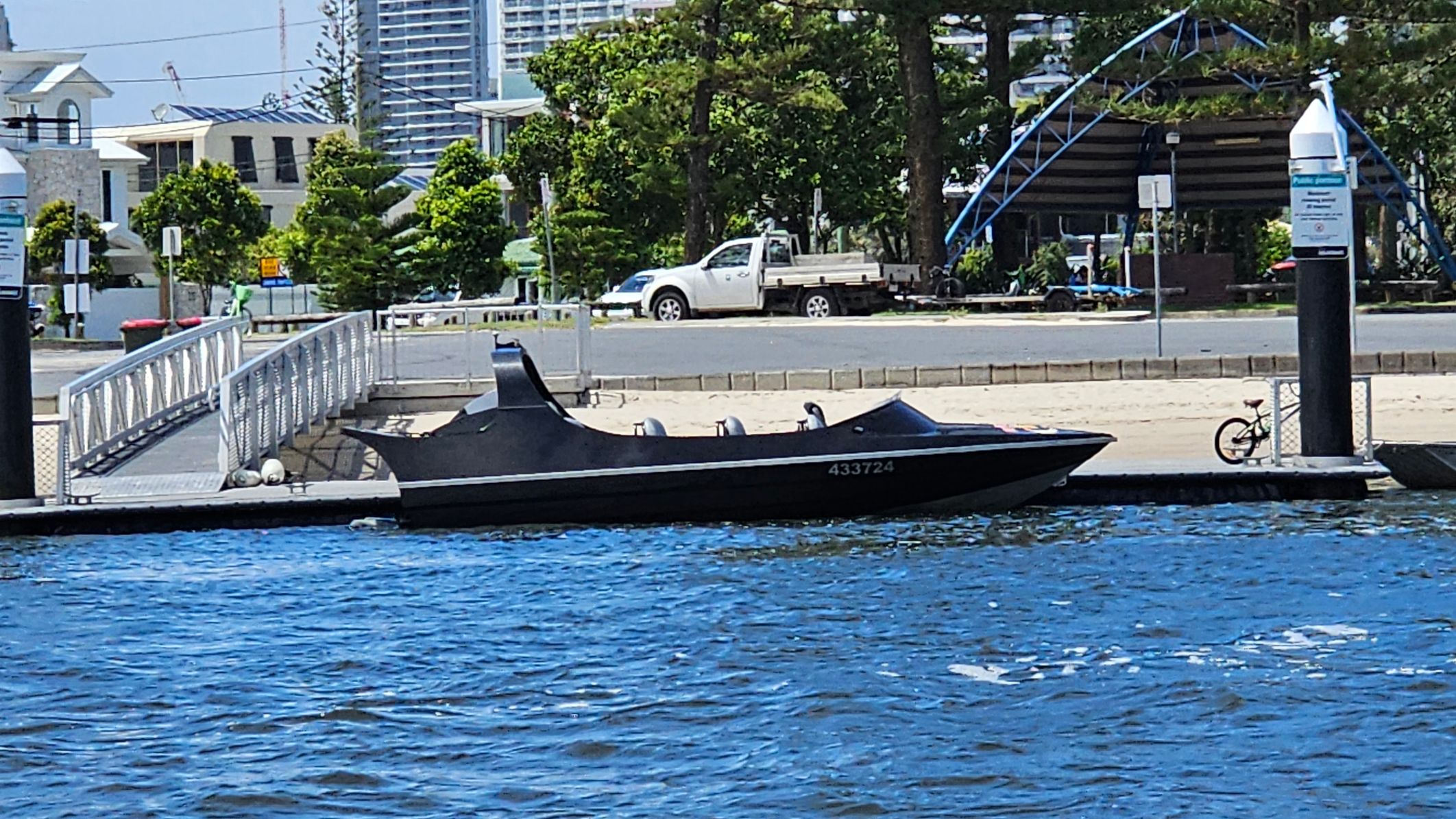 A sleek black boat is moored at a dock on a body of water. In the background, there is a white pickup truck parked on a road, with several trees and buildings visible. A metal ramp leads down to the dock, and a bicycle is parked near a post. A large, blue, arched structure is partially visible behind the trees. The water in the foreground is a deep blue with small ripples. A sleek black boat is moored at a dock on a body of water. In the background, there is a white pickup truck parked on a road, with several trees and buildings visible. A metal ramp leads down to the dock, and a bicycle is parked near a post. A large, blue, arched structure is partially visible behind the trees. The water in the foreground is a deep blue with small ripples.
