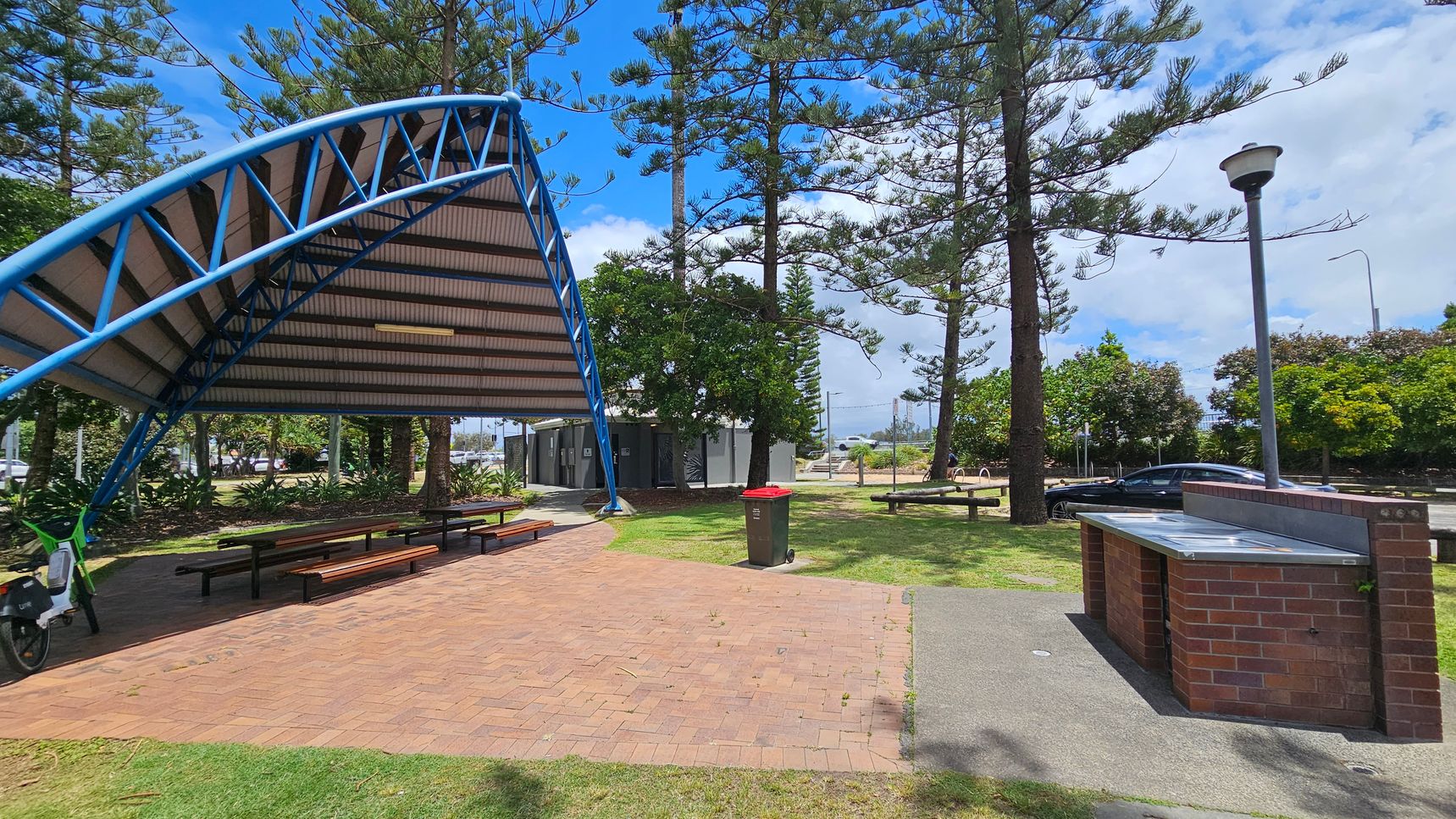 A park scene with a covered picnic area on the left, featuring a blue metal frame and a corrugated roof. Beneath the shelter are several picnic tables. A bicycle is parked nearby. In the centre, there is a brick path leading to a bin with a red lid. On the right, a brick barbecue stand is visible, along with a lamp post. Tall trees and bushes are scattered throughout the grassy area, and a car is parked in the background. A park scene with a covered picnic area on the left, featuring a blue metal frame and a corrugated roof. Beneath the shelter are several picnic tables. A bicycle is parked nearby. In the centre, there is a brick path leading to a bin with a red lid. On the right, a brick barbecue stand is visible, along with a lamp post. Tall trees and bushes are scattered throughout the grassy area, and a car is parked in the background.