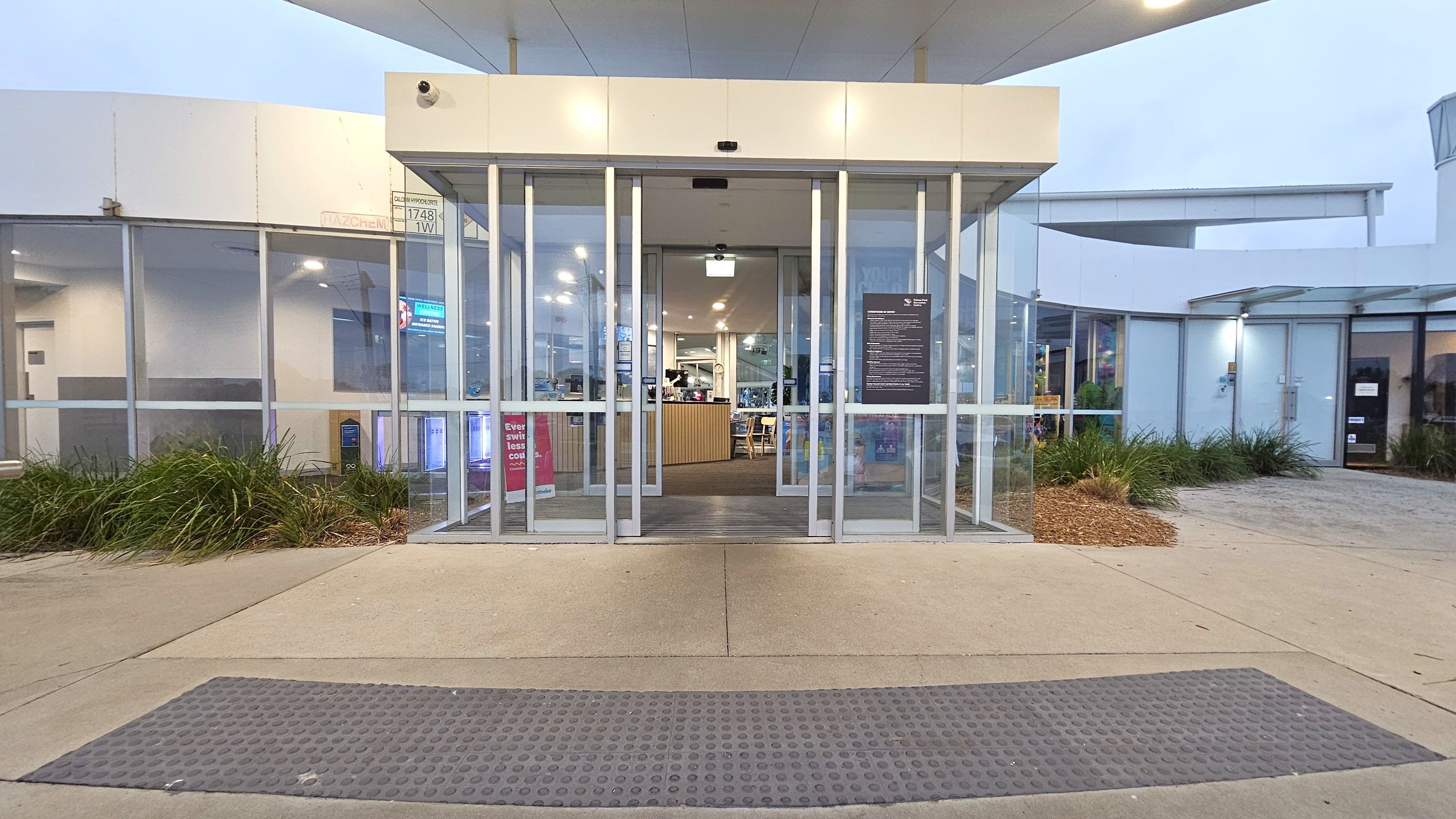 A modern glass entrance with automatic sliding doors is centred in the image. Inside, a reception area with a wooden counter and seating is visible. On either side of the entrance, there are large glass panels and some signage. The foreground features a textured rubber mat on a concrete path. Surrounding the entrance, there are landscaped areas with green plants and mulch. The ceiling above the entrance is white with recessed lighting.