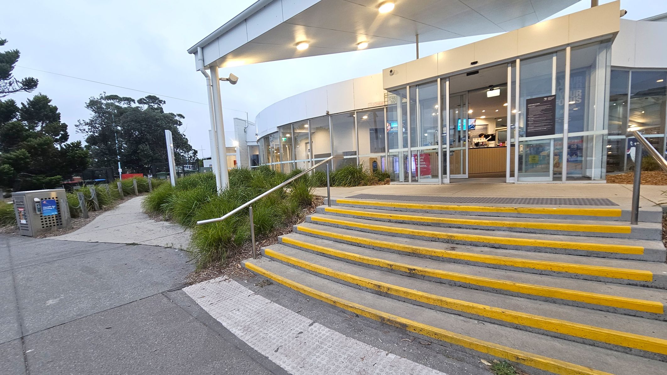 A modern building entrance with large glass doors is on the right, featuring a set of steps with yellow edges leading up to it. The ceiling above the entrance has recessed lighting. On the left, a pathway lined with green shrubs and trees leads away from the building. A metal bin is visible near the path. The ground is a mix of concrete and textured surfaces, and there are metal railings along the steps.