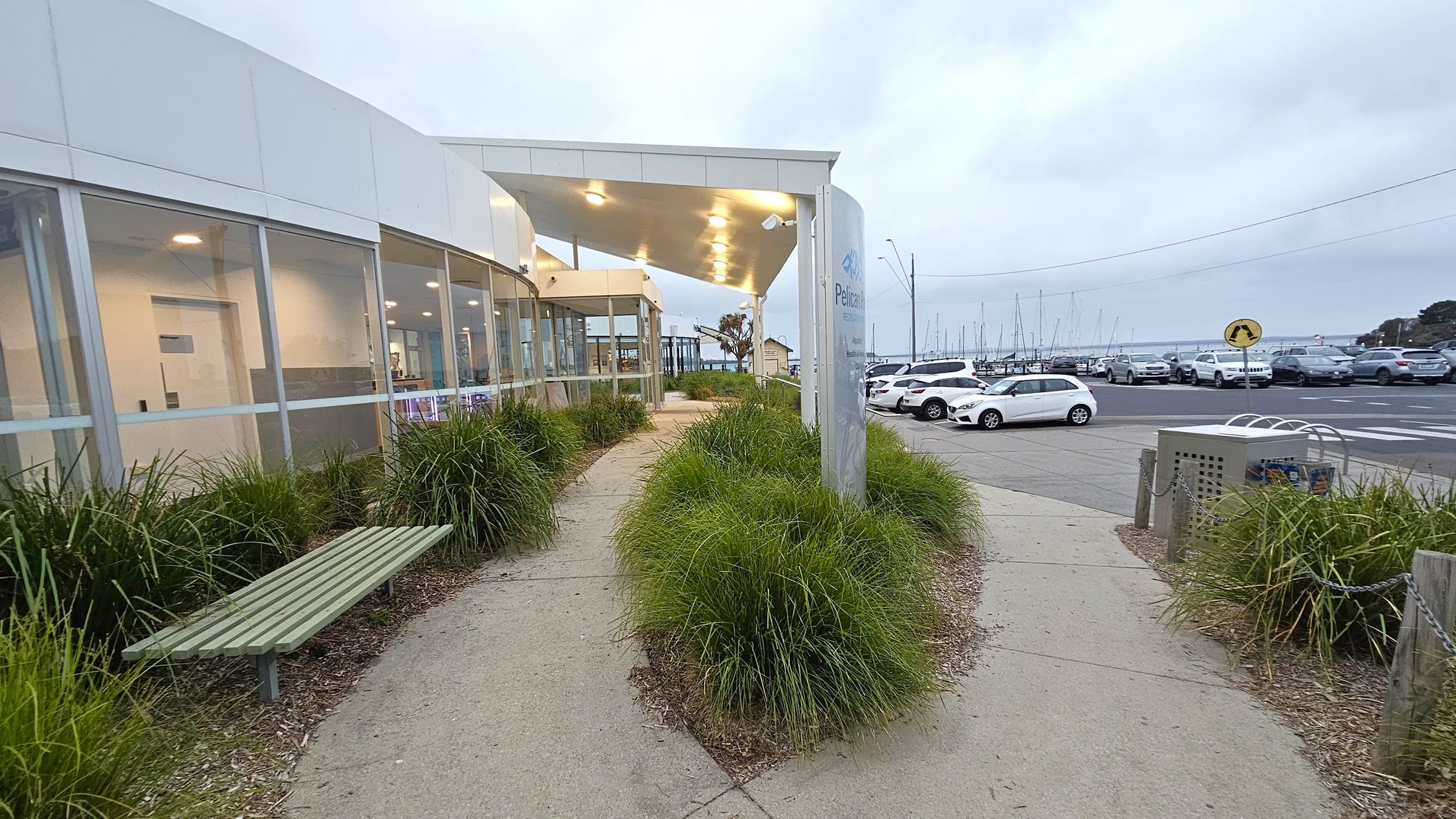 A modern building with large glass windows on the left, featuring a green bench and landscaped plants along a curved path. The path leads to a covered entrance with lights. On the right, a car park with several vehicles and a view of sailboats in the distance. A signpost and a small utility box are near the path. The area is well-maintained with neatly trimmed grass and shrubs.