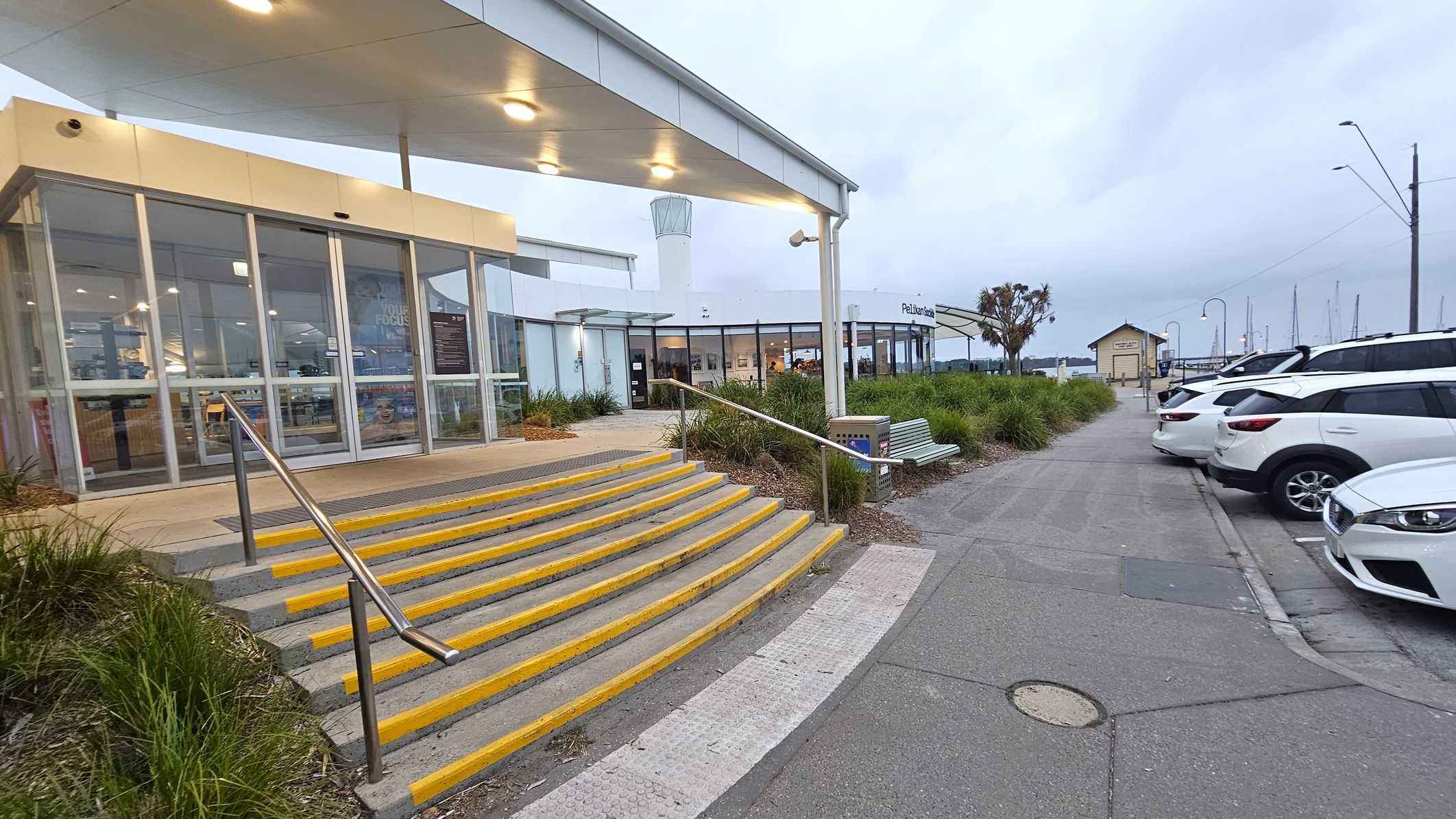 On the left, there is a building with large glass windows and a set of steps with yellow edges leading up to it. A metal handrail runs alongside the steps. The building has a covered entrance with lights on the ceiling. In the middle, there is a pathway lined with green plants and a bench. On the right, several white cars are parked in a row. In the background, there are more buildings, a small structure, and masts from boats, suggesting a marina setting.