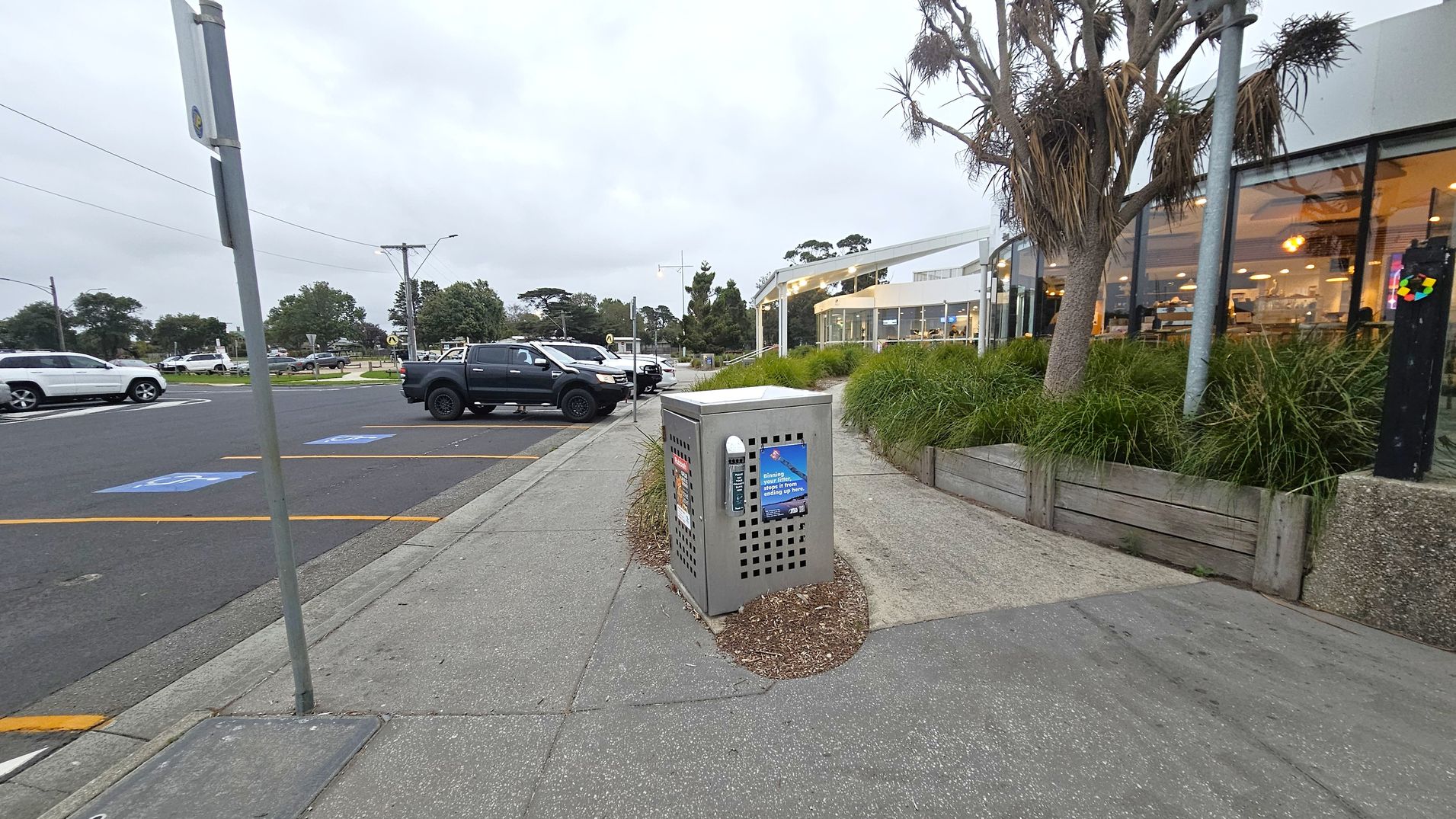 A pavement with a grey utility box in the centre, surrounded by a small patch of mulch. On the right, there's a building with large glass windows, behind which are visible tables and chairs. A tree and some tall grass are planted in a raised bed next to the building. On the left, a car park with several vehicles, including a white SUV and a dark pickup truck. A signpost is visible near the kerb. The ground is paved with concrete, and the area appears to be a public space.