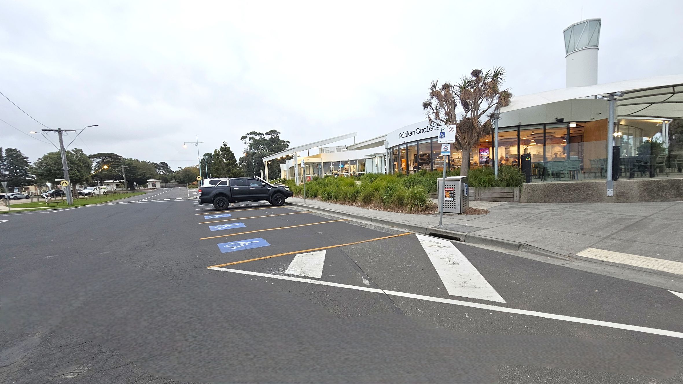 A car park with designated disabled parking spaces marked in blue is on the left. A black vehicle is parked in one of the spaces. On the right, a modern building with large glass windows and a sign reading "Pelikan Societ" is visible. The building is surrounded by landscaped greenery, including small trees and shrubs. A rubbish bin is placed near the entrance. The pavement is grey, and a tall structure with a glass top is in the background.