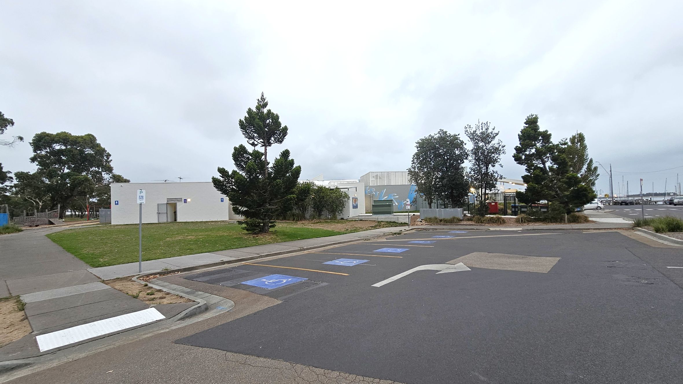 A paved parking area with marked disabled parking spaces and directional arrows in the foreground. On the left, a small white building with a door and signage is surrounded by grass and trees. In the middle, more trees and a mural on a building wall are visible. The background shows a marina with boats and masts. The area is bordered by a footpath and some low vegetation.