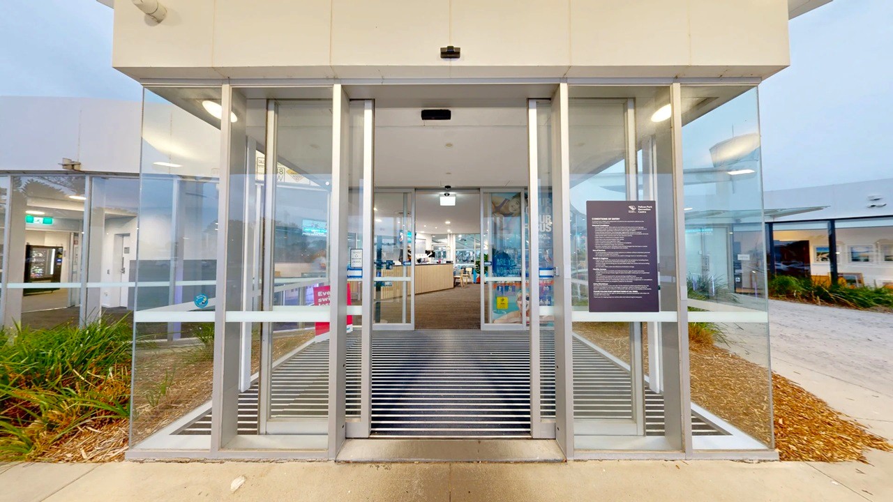 Glass entrance with automatic sliding doors, framed by metal. Inside, a reception area with a desk and various items is visible. To the left, a corridor leads to other rooms, with an emergency exit sign above. On the right, a large sign with text is mounted on the glass. The exterior has a concrete path and landscaped areas with plants. The ceiling inside is white with recessed lighting, and the floor outside is concrete with a metal grate at the entrance.