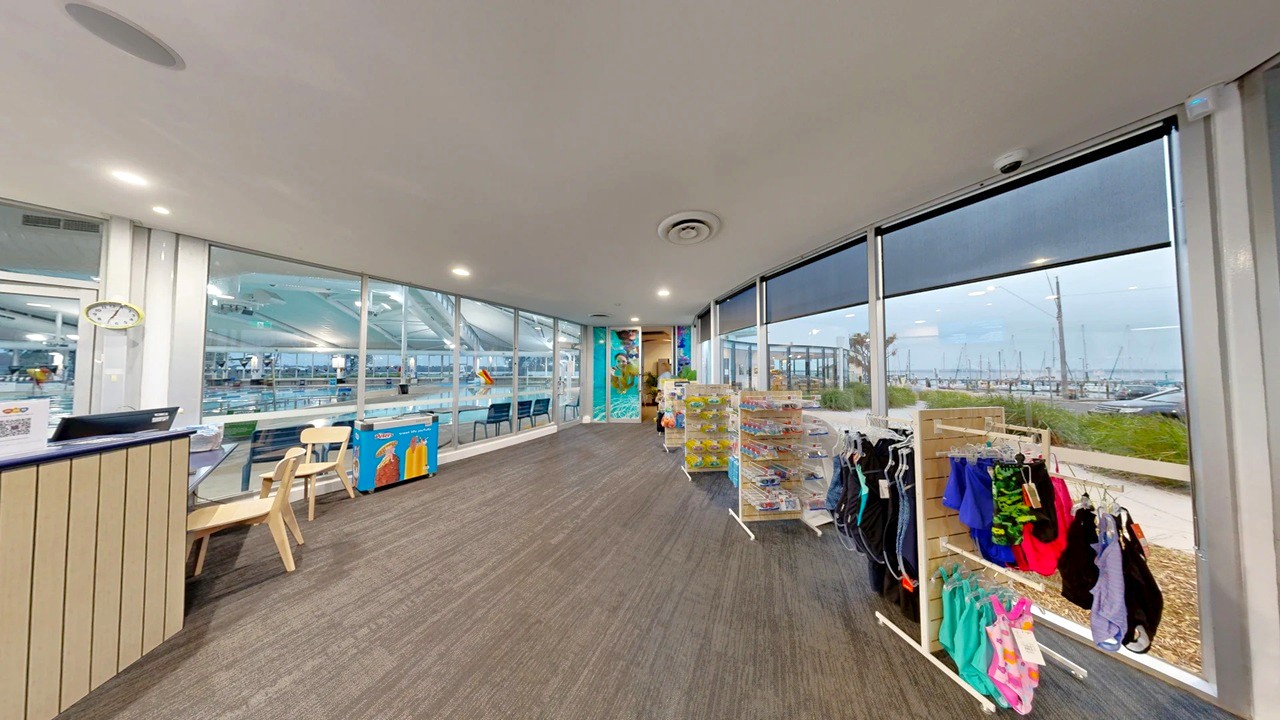 A spacious indoor area with large windows on the right, offering a view of a marina with boats. On the left, there's a reception desk with a clock above it and a couple of chairs. In the centre, there are display racks with swimwear and accessories. The floor is covered with dark wood-like material, and the ceiling is white with recessed lighting. The background features a colourful mural and more seating near the windows.