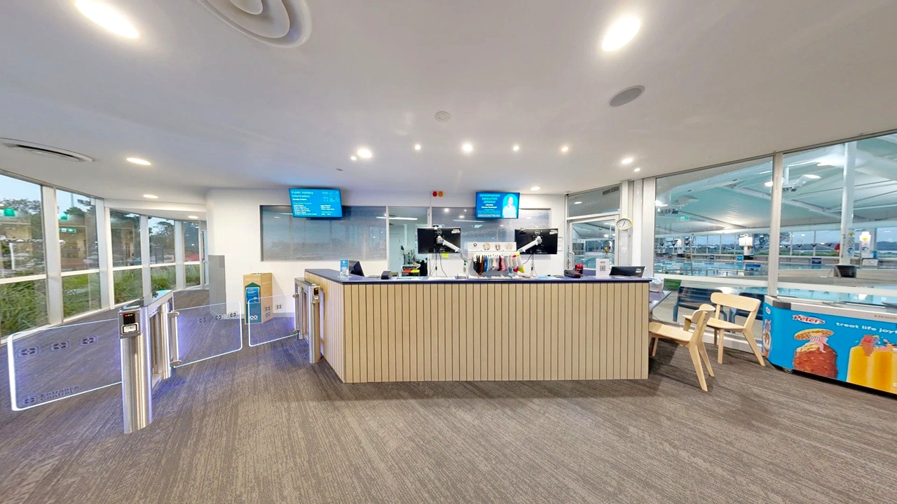 A modern reception area with a wooden counter in the centre, equipped with computers and microphones. Two digital screens are mounted on the wall behind the counter. On the right, there are wooden chairs and a display with snacks and drinks. The left side features a series of turnstiles leading to a glass wall with a view of greenery outside. The floor is carpeted, and the ceiling has recessed lighting.