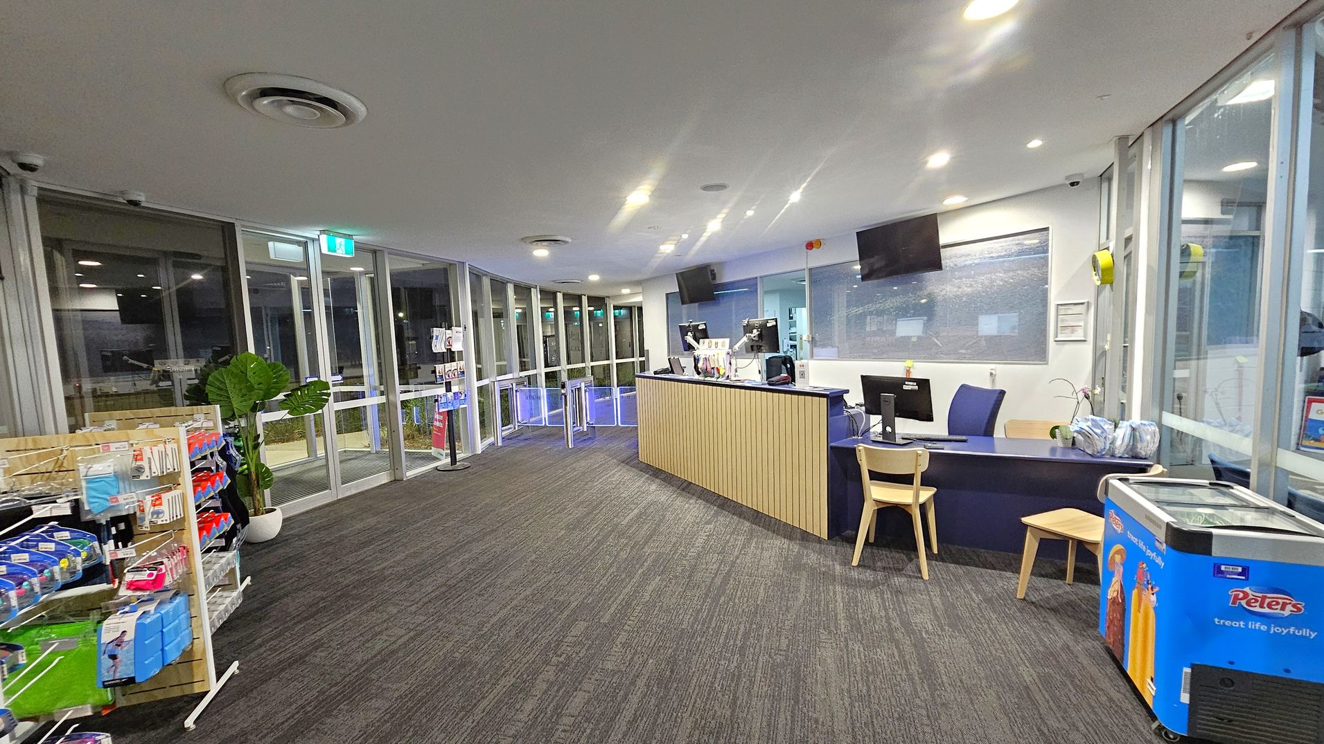 A reception area with a modern design. On the left, there are glass doors and windows, a plant, and a display rack with various items. In the centre, a wooden reception desk with computers and chairs. On the right, a blue ice cream freezer with "Peters" branding and more glass windows. The floor is carpeted with a patterned design, and the ceiling is white with recessed lighting.