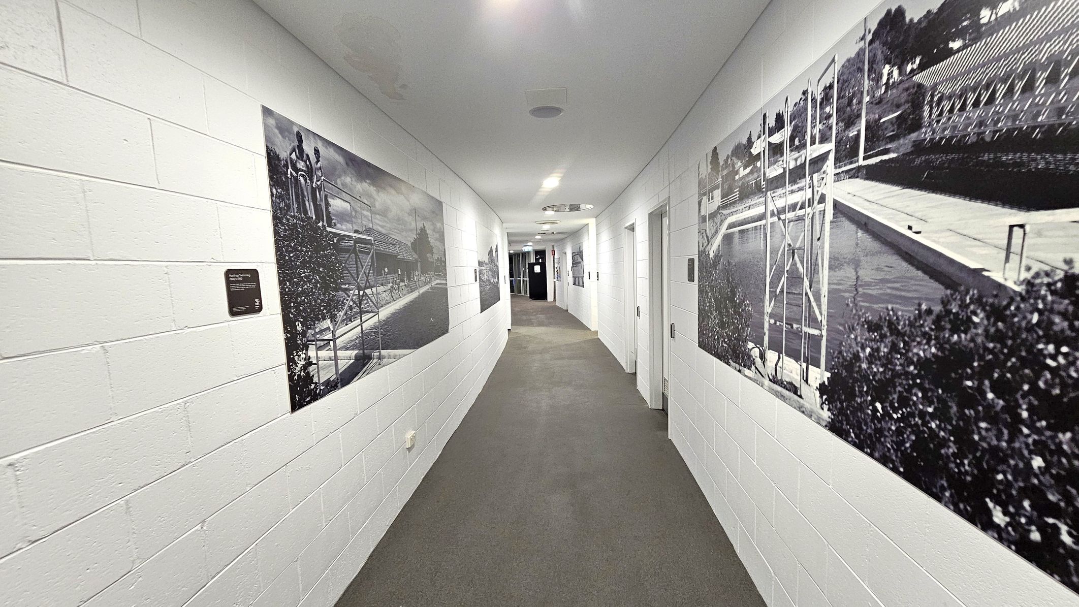 A long corridor with white brick walls and a grey carpeted floor. On the left wall, there are large black and white photographs depicting scenes of a swimming pool and people. The right wall also features similar large photographs of a pool area. Several doors are visible on the right side, leading to different rooms. The ceiling is white with recessed lighting, and the corridor curves slightly towards the end, where a door is visible.