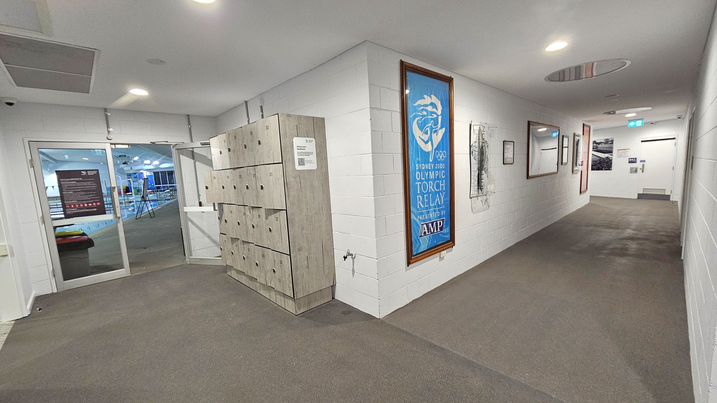 A hallway with white brick walls and a brown and grey carpet. On the left, there are wooden lockers and a glass door leading to a swimming pool area. The right wall features framed posters, including one for the Sydney 2000 Olympic Torch Relay. The ceiling is white with recessed lighting, and there is a circular vent. At the end of the hallway, there is a door with a sign and an emergency exit light above it.