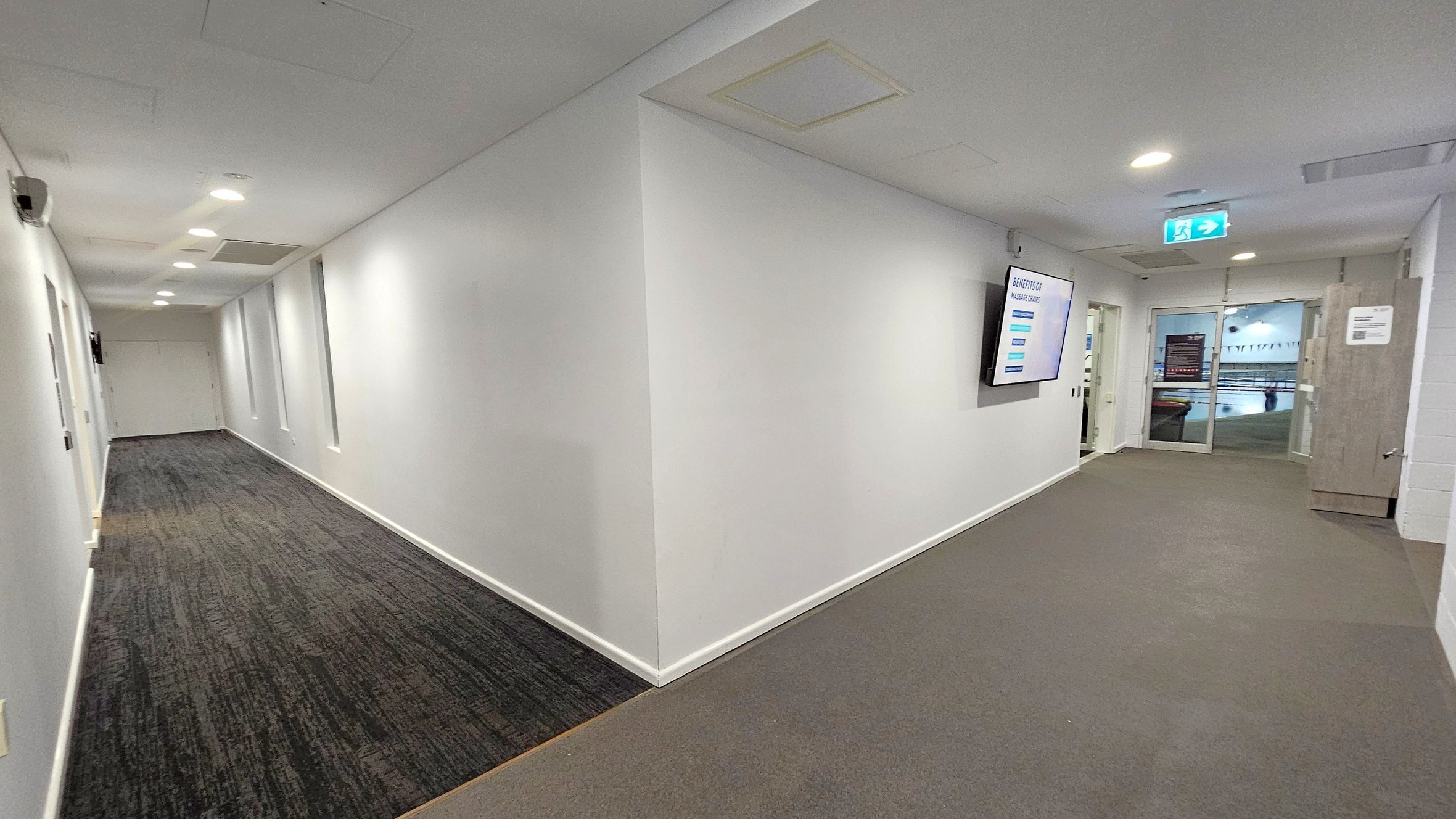 A hallway with white walls and a grey carpeted floor. On the left, the corridor extends with several ceiling lights and a dark carpet. On the right, a wall-mounted screen displays information. Further down, a glass door leads to another area, possibly a pool, visible through the glass. The ceiling is white with recessed lighting and ventilation panels.