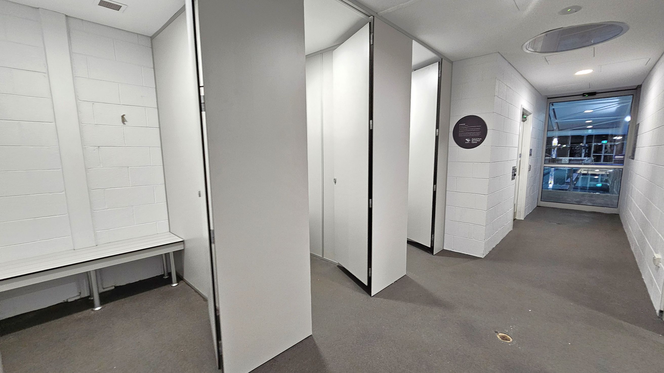 A corridor with white brick walls and a grey carpeted floor. On the left, there is a white bench with metal legs. In the centre, several open doors lead to changing cubicles. On the right, a circular sign is mounted on the wall. At the end of the corridor, a large window reveals a view of an indoor area with visible lights and structures. The ceiling is white with recessed lighting and a circular vent.