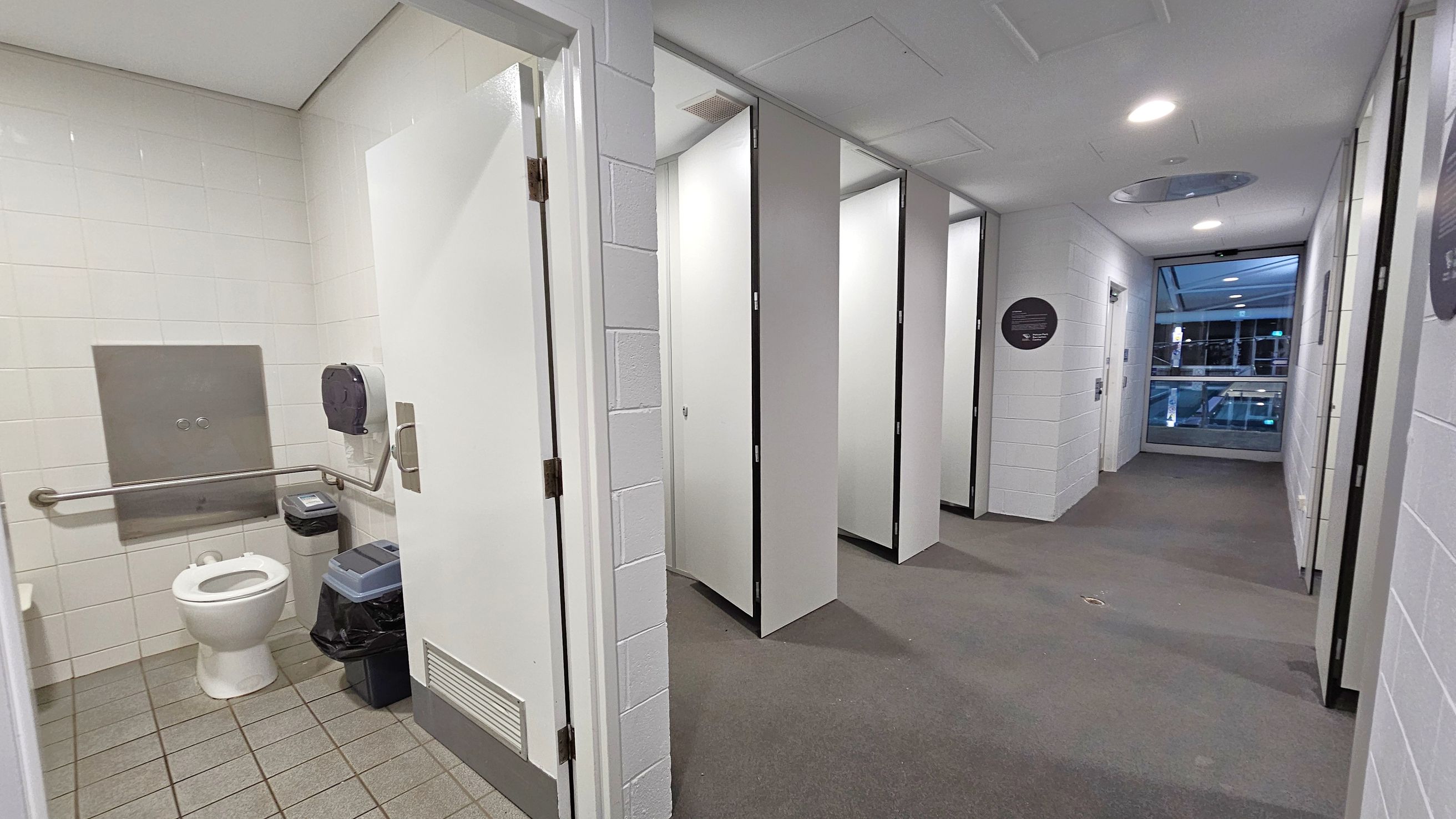 A public restroom with a tiled floor and white walls. On the left, an open stall reveals a toilet, grab bars, a toilet paper dispenser, and a bin. The middle area shows a row of closed stall doors. The right side features a corridor with a grey carpet leading to a windowed area. The ceiling is white with recessed lighting.