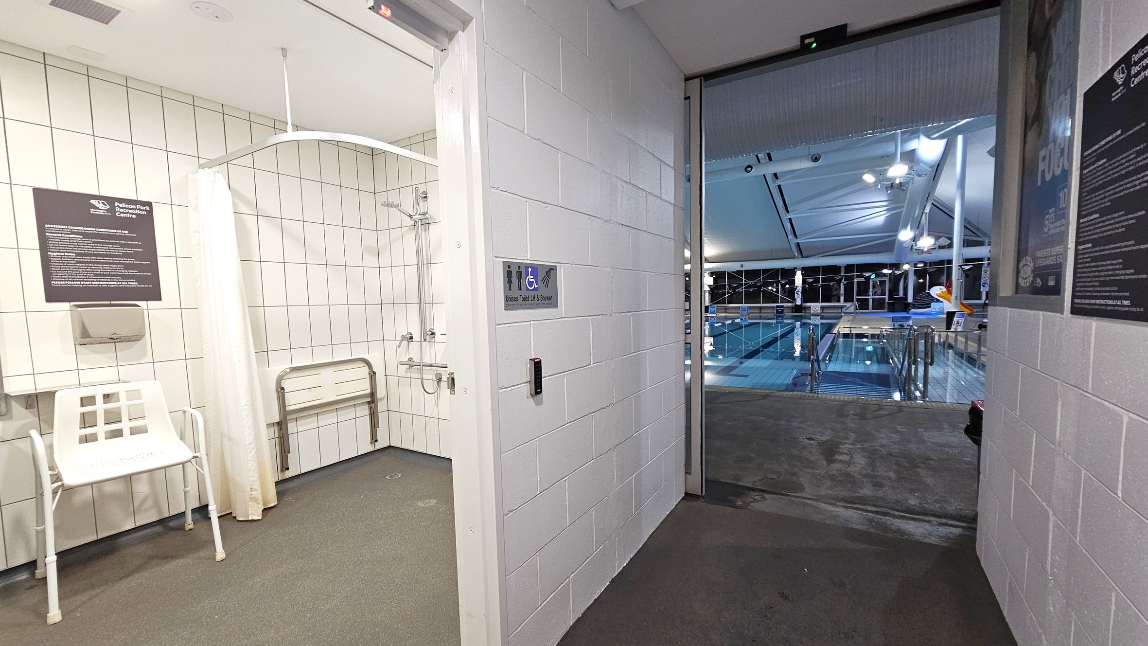 On the left, there is a tiled room with a shower, a white chair, and a folding bench. A sign and a paper towel dispenser are mounted on the wall. In the centre, a door leads to a pool area. The wall beside the door has a sign indicating a unisex toilet and shower. On the right, the pool area is visible with a large swimming pool, metal railings, and a high ceiling with lights. The floor is a mix of tiles and concrete.
