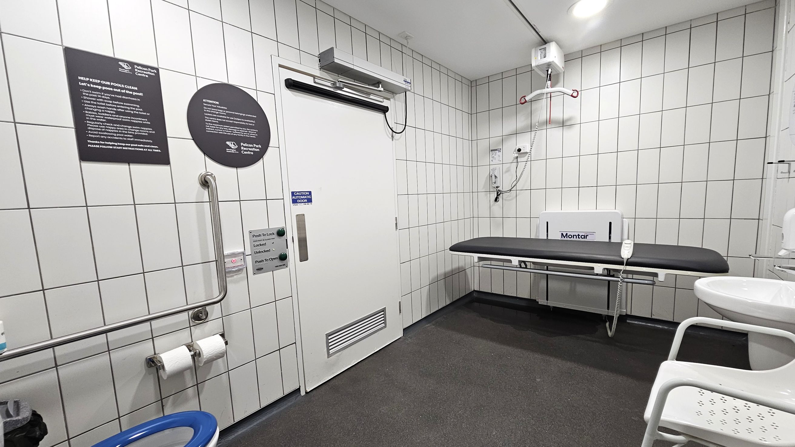 A tiled room with a white automatic door on the left, featuring a push-to-lock mechanism and informational signs. Below the signs, there is a grab rail and two toilet paper rolls. On the right, a black changing table is mounted on the wall, with a hoist system above it. A white sink with a chair is partially visible in the foreground. The floor is dark and the ceiling is white with recessed lighting.