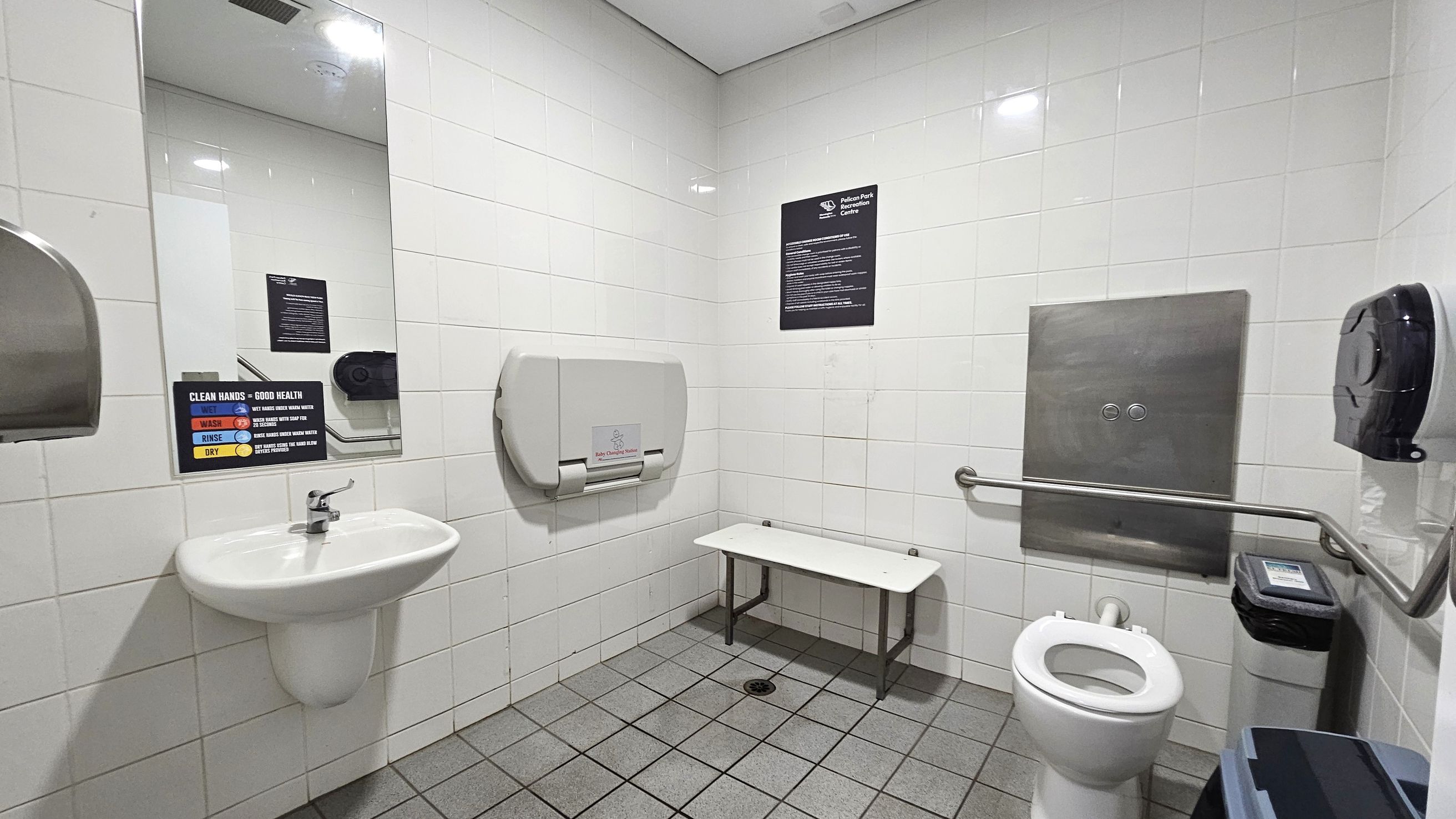 A public restroom with white tiled walls and a grey tiled floor. On the left, a white sink with a mirror above it and a handwashing instruction sign. A hand dryer is mounted on the wall. In the centre, a baby changing station is attached to the wall above a small bench. On the right, a toilet with a grab rail beside it, a toilet paper dispenser, and a waste bin. A stainless steel panel is mounted on the wall above the toilet. The ceiling is white with recessed lighting.