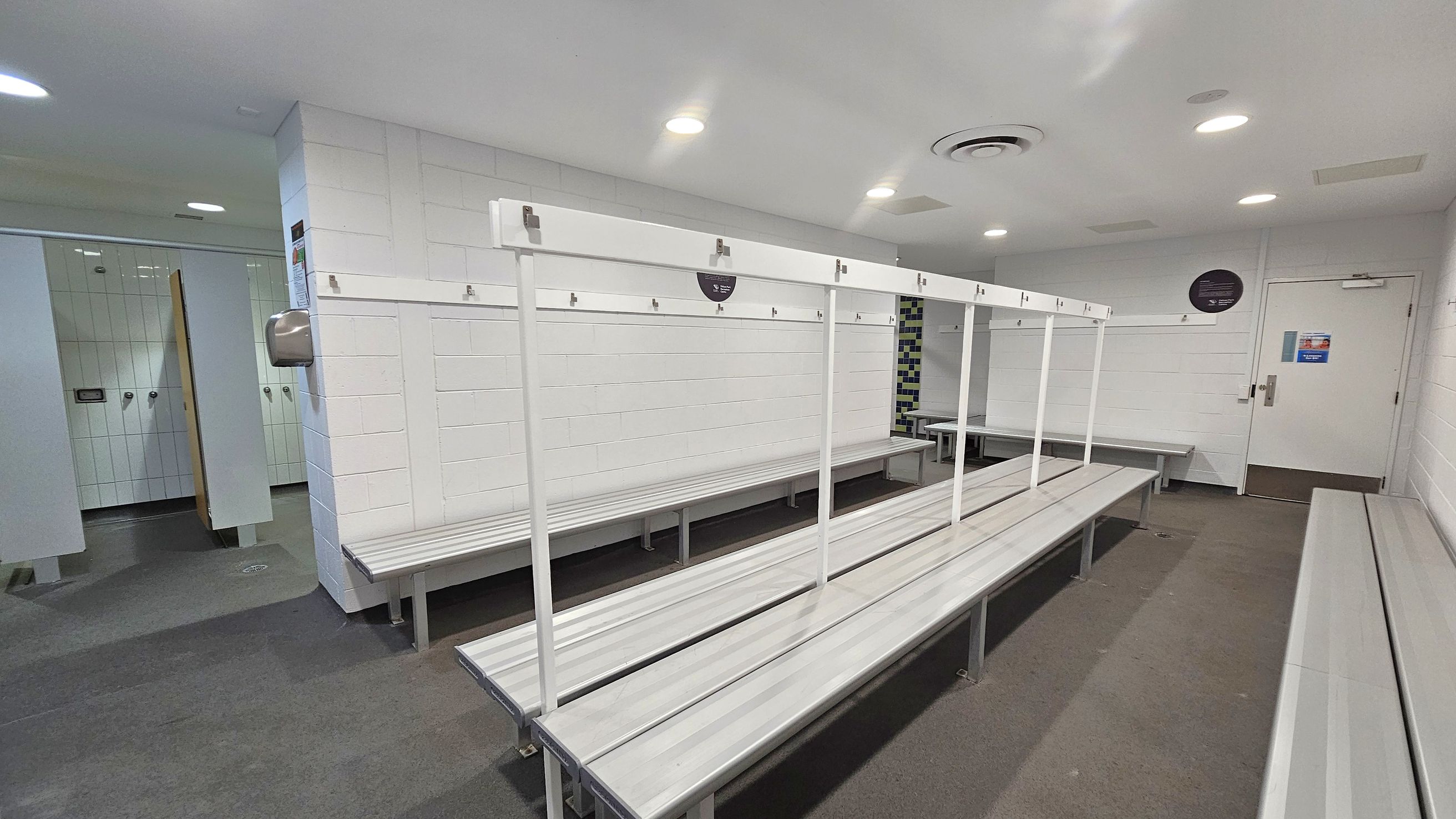 A locker room with white tiled walls and grey flooring. On the left, there are shower cubicles with open doors. In the centre, long metal benches with hooks above them run parallel to each other. On the right, a white door with a sign and a notice board is visible. The ceiling is white with recessed lighting and an air vent.