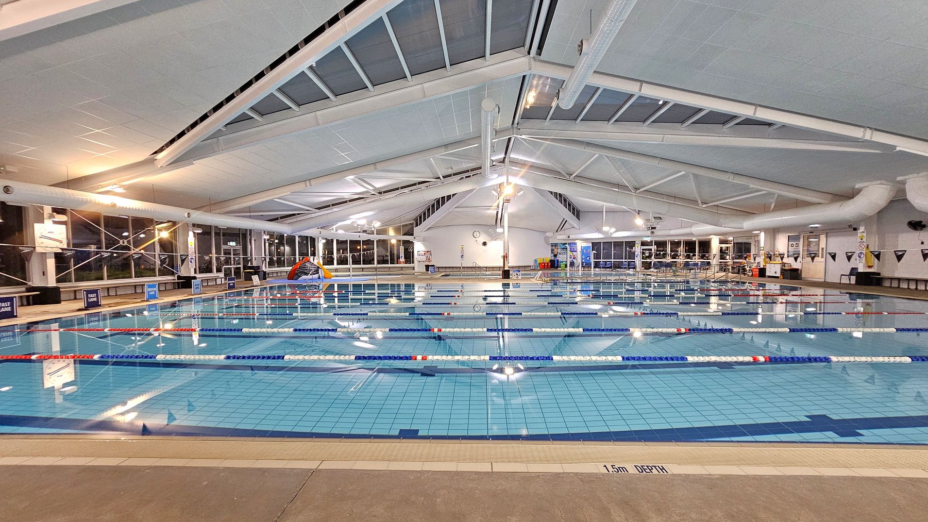 An indoor swimming pool with multiple lanes, separated by blue and white lane dividers. The pool area is well-lit, with large windows on the left side allowing natural light. The ceiling is high and sloped, featuring white beams and skylights. On the right, there are seating areas and a few doors. The pool deck is tiled, and a sign indicates a depth of 1.5 metres. In the background, there are colourful pool toys and a clock on the wall.