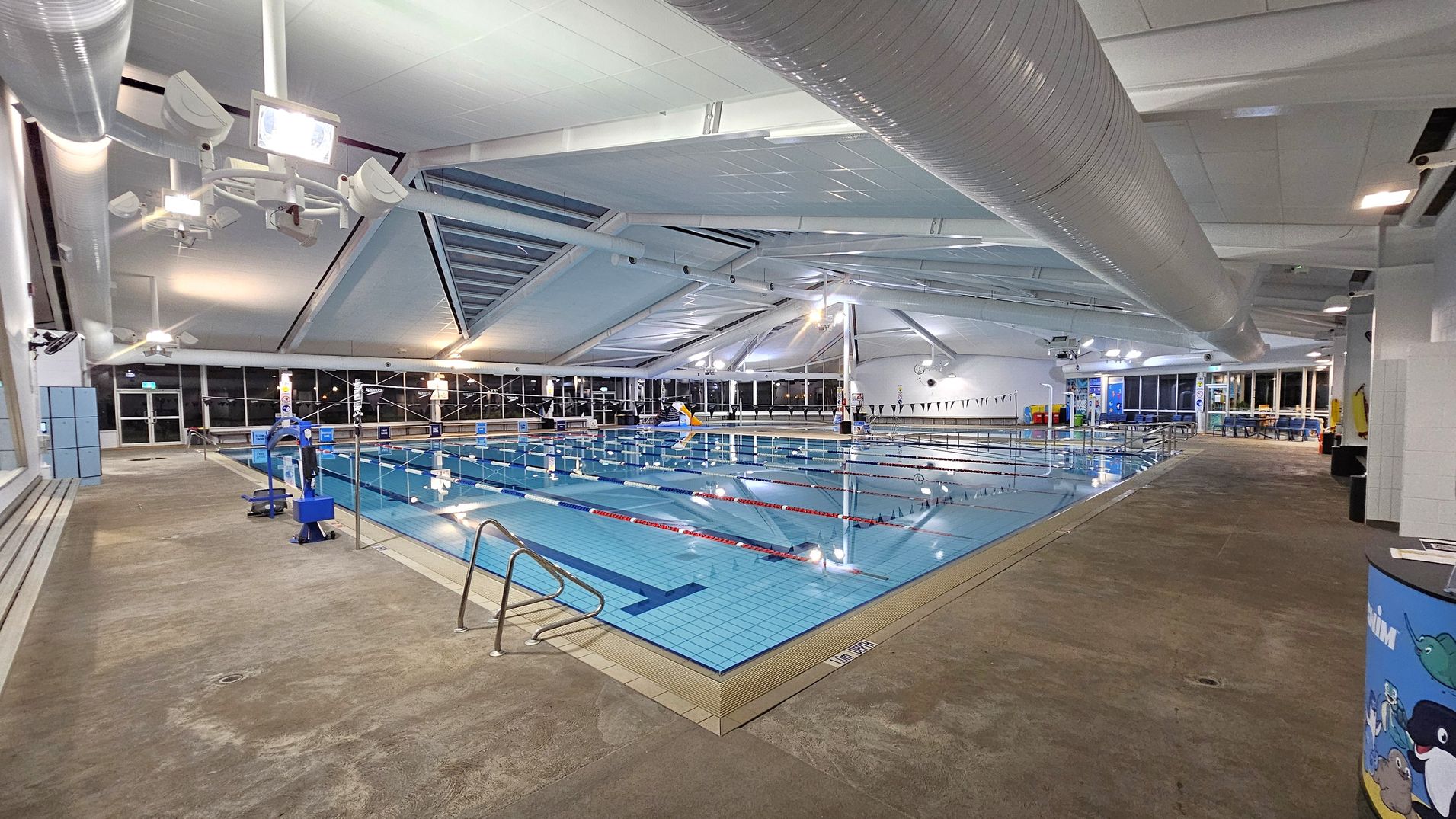 An indoor swimming pool with clear blue water is in the centre, divided into lanes by red and blue ropes. On the left, there are blue lockers and a bench along the wall. The right side features a wide walkway with a colourful cylindrical bin. The ceiling is high with large white ducts and bright lighting. The floor is a textured, non-slip surface. Large windows line the background, allowing natural light to enter.